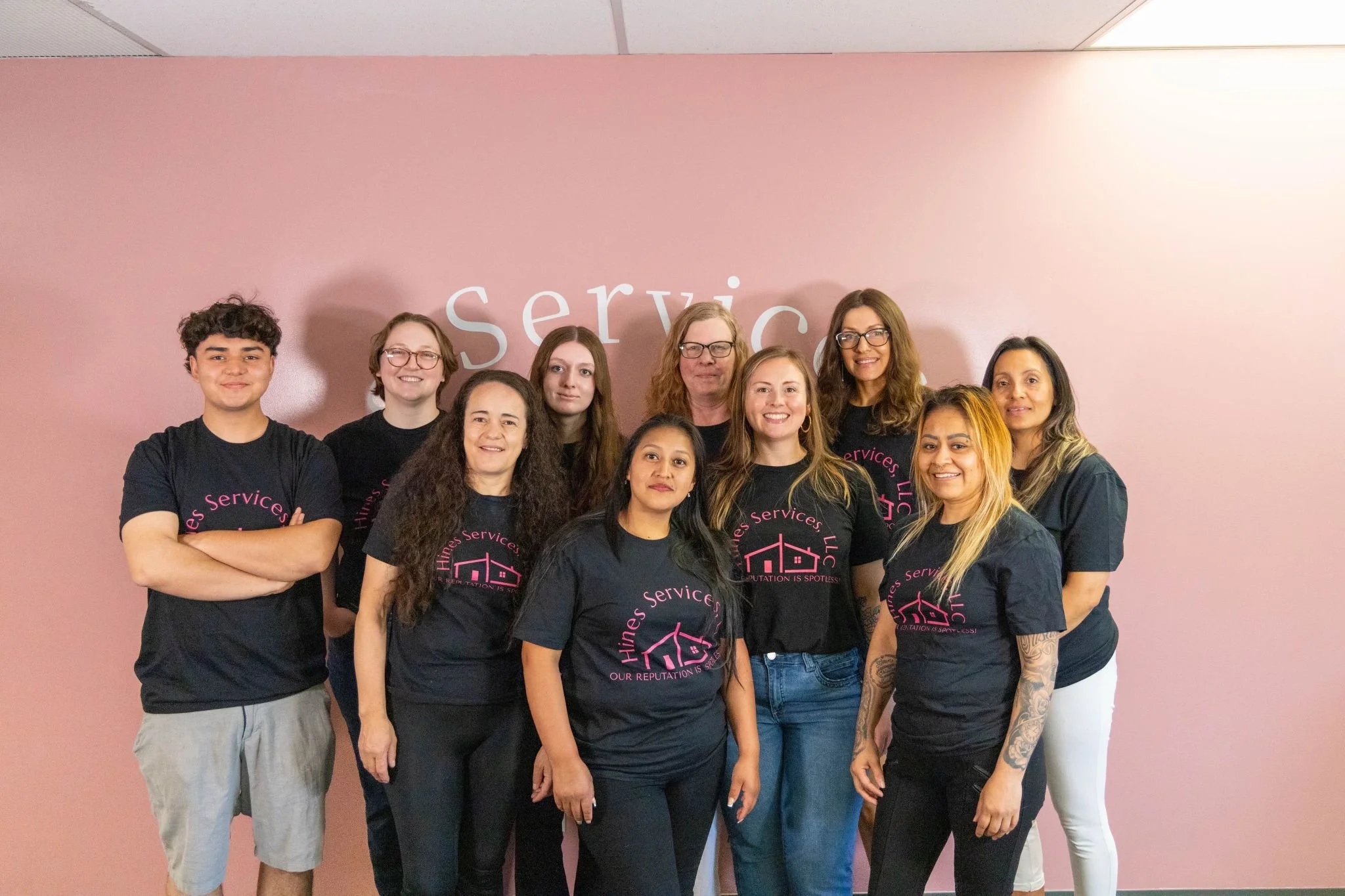 Group of diverse women and one young man standing together in front of a pink wall, all wearing matching black T-shirts with pink lettering and graphics for an organization or event.