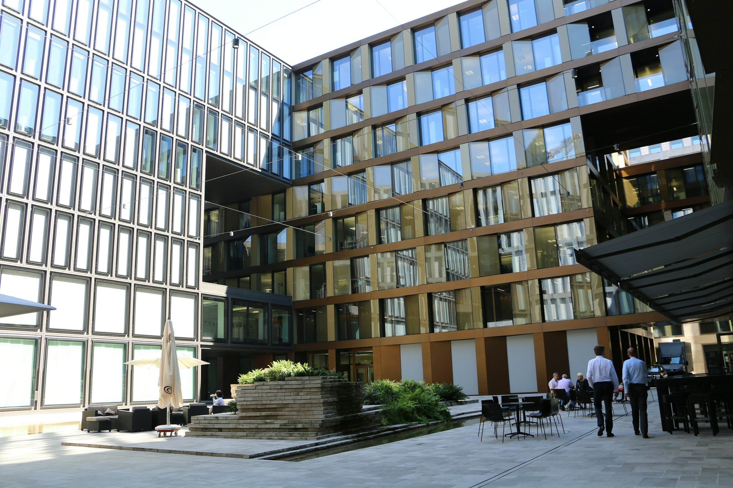 Modern office building with glass windows and a courtyard featuring outdoor seating, plants, and people walking and socializing.