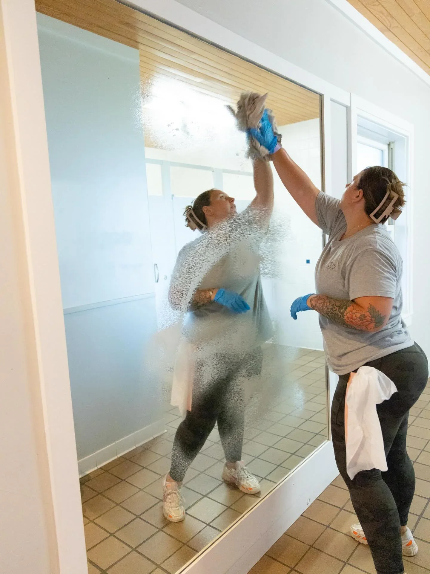 Two women cleaning a large glass window from the inside. They are wearing gray t-shirts, gloves, and hair covers, with cleaning cloths in hand.