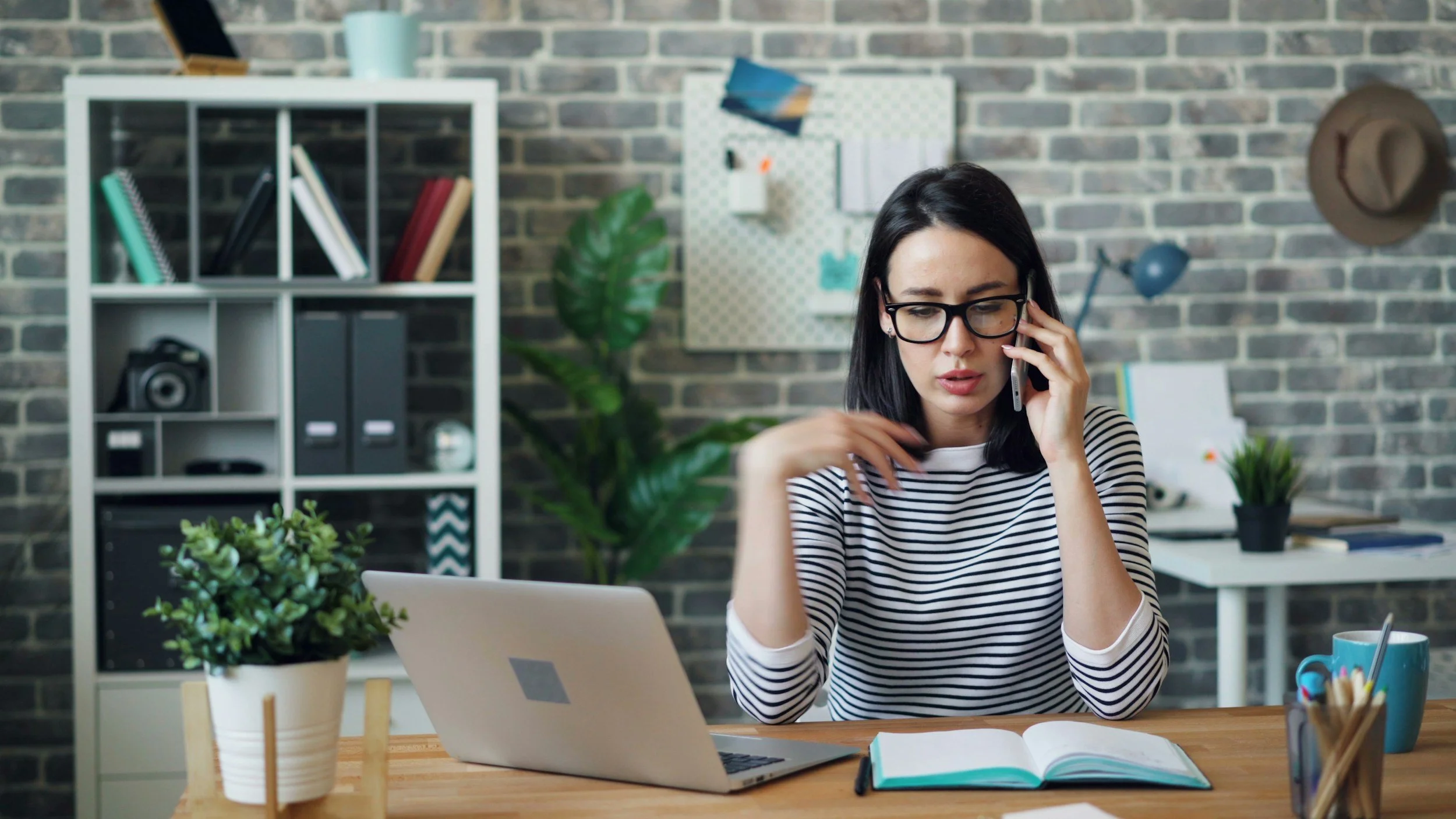 A woman with glasses and dark hair working at her desk, talking on the phone, with a laptop, open notebook, and office supplies. Behind her are shelves with books, binders, and a camera, as well as a plant and a corkboard on a brick wall.
