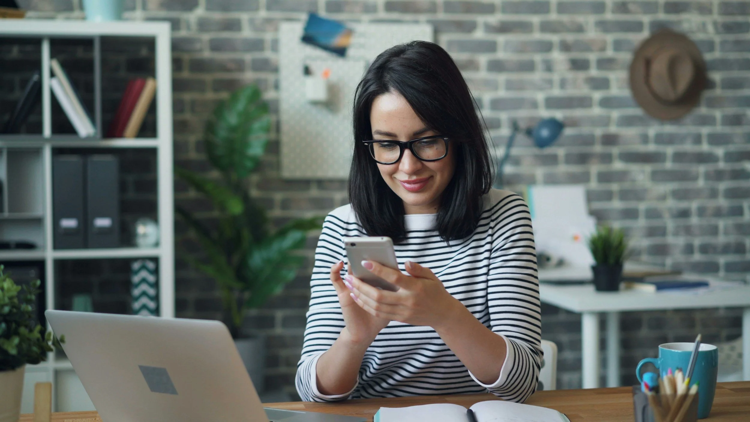 A woman with black hair and glasses wearing a black and white striped shirt, smiling while looking at her smartphone in a modern office or workspace setting.