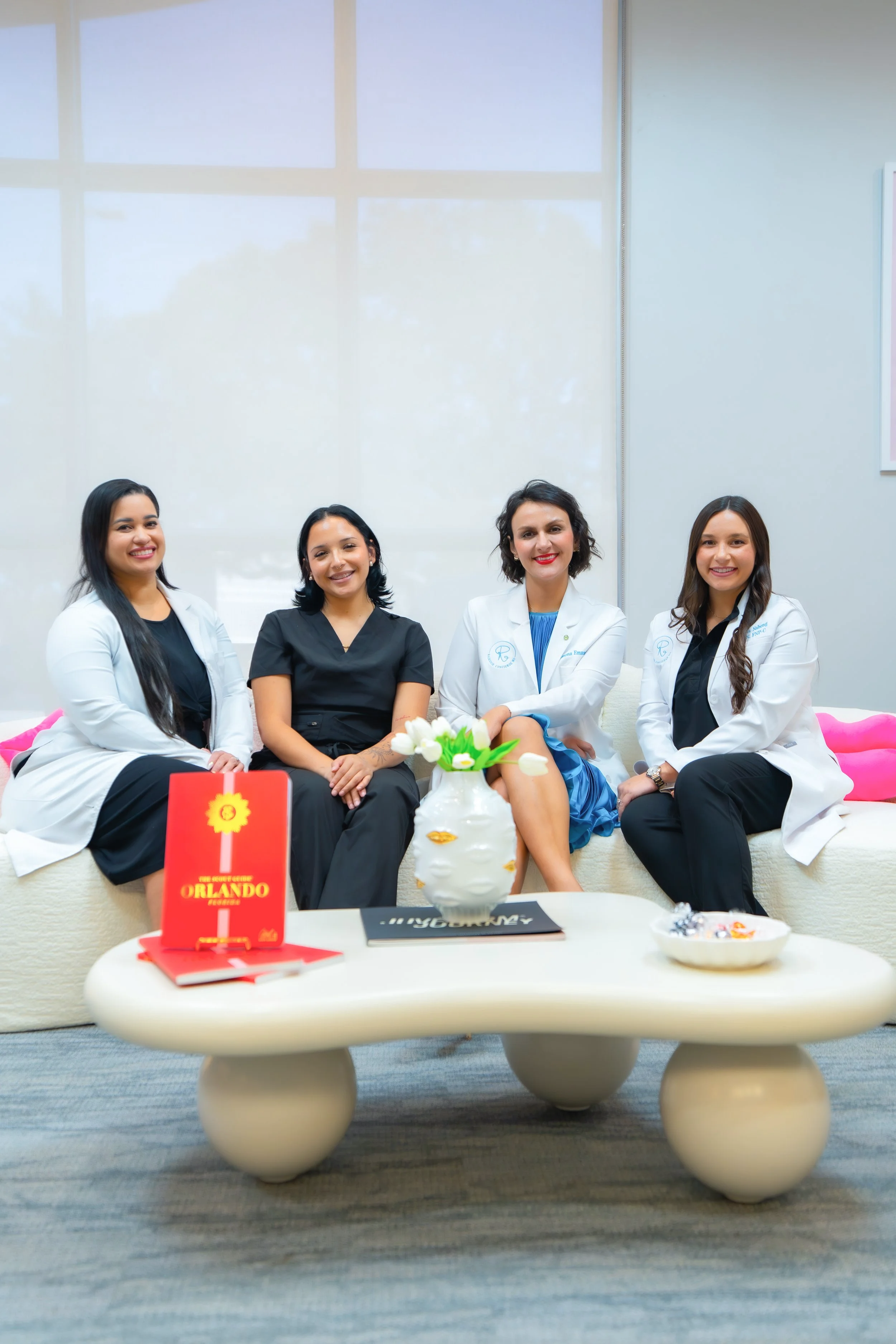 Four women sitting on a white couch in an office or clinic lounge, smiling for the camera. They are dressed in medical scrubs and white coats, with a coffee table in front of them holding a red book or brochure, a vase with white tulips, and a bowl of candy.
