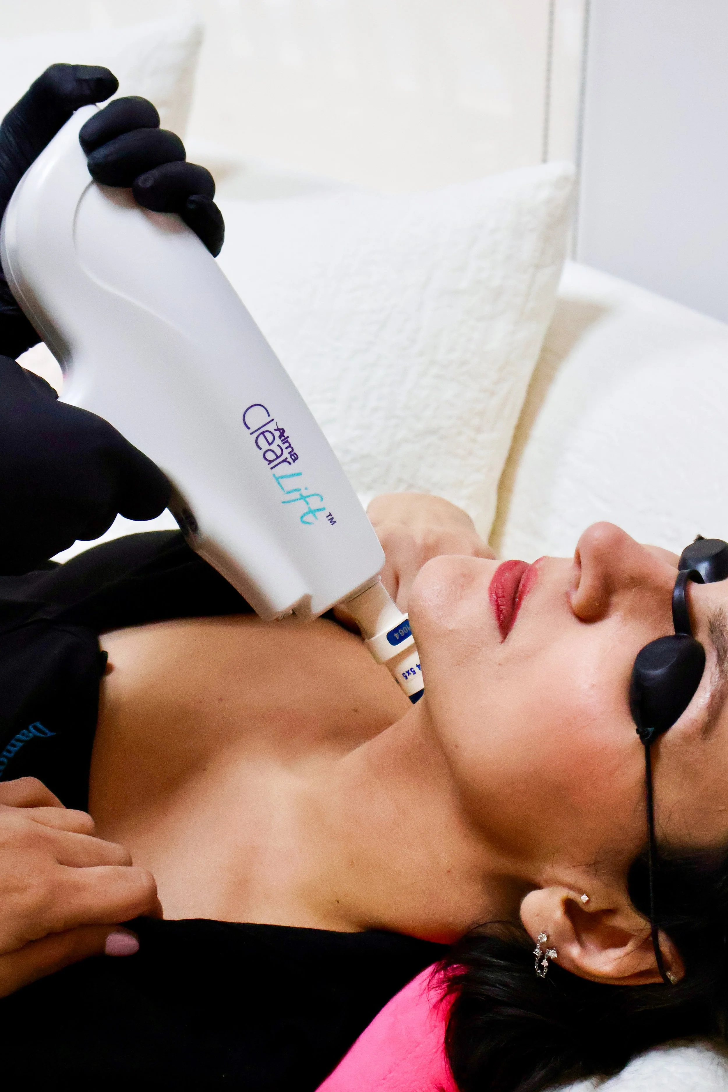A woman lying on a bed receiving a cosmetic treatment with a handheld device, wearing protective glasses, and holding her chest with one hand.
