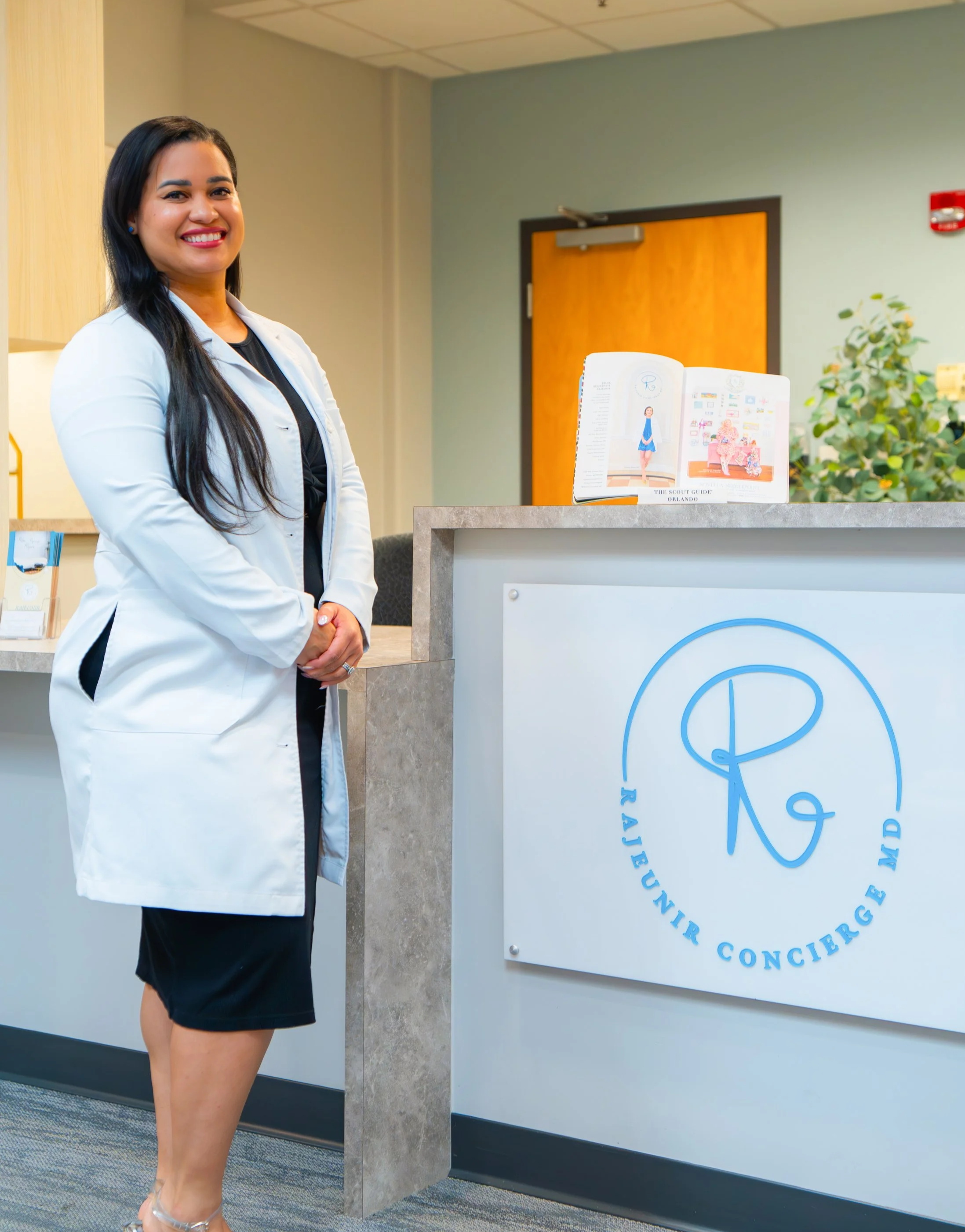 A smiling woman in a white coat standing behind a reception desk with a sign that reads 'R aims at your overnight care MD.'