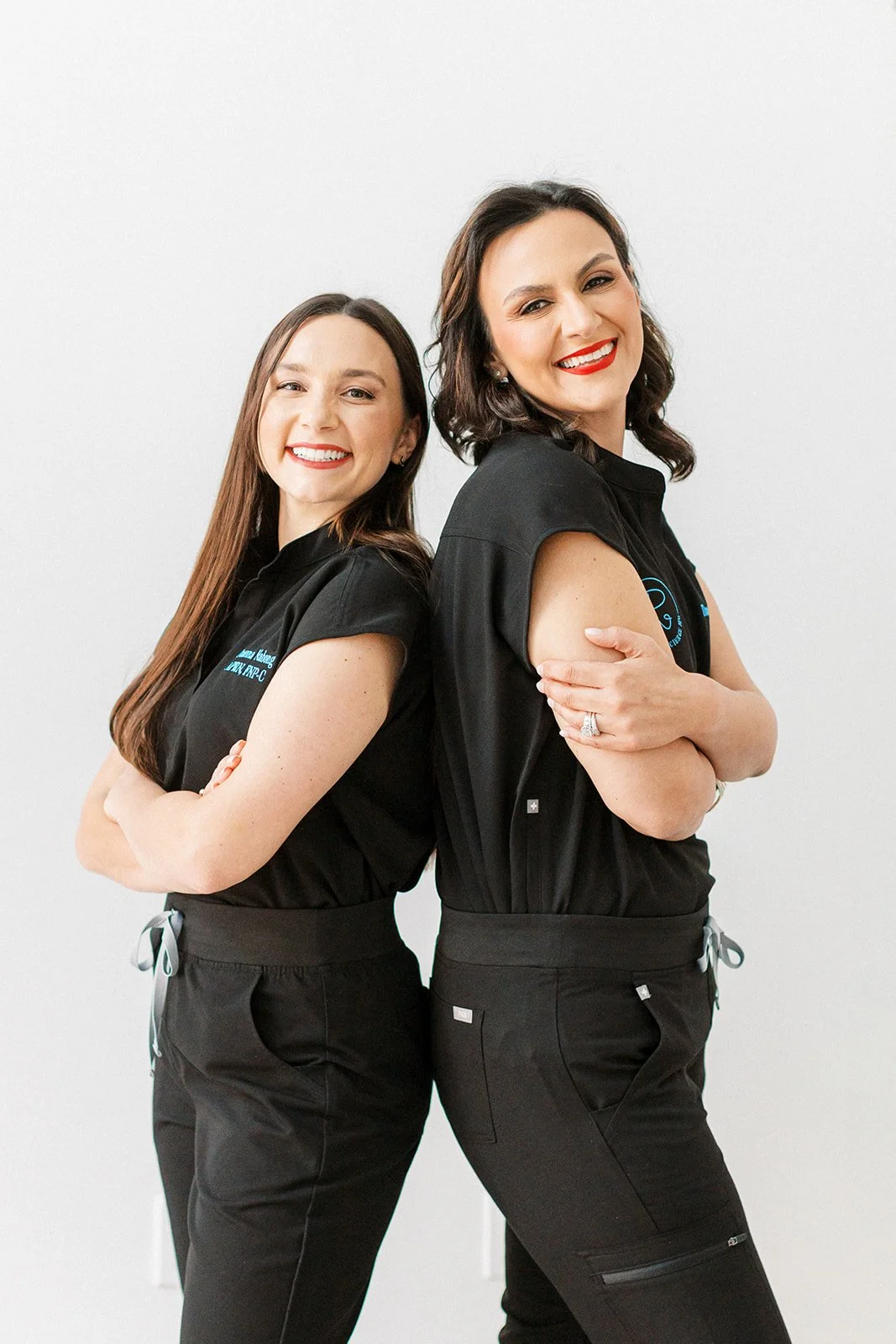 Two women standing back to back with arms crossed, smiling, wearing black uniforms, against a plain white background.