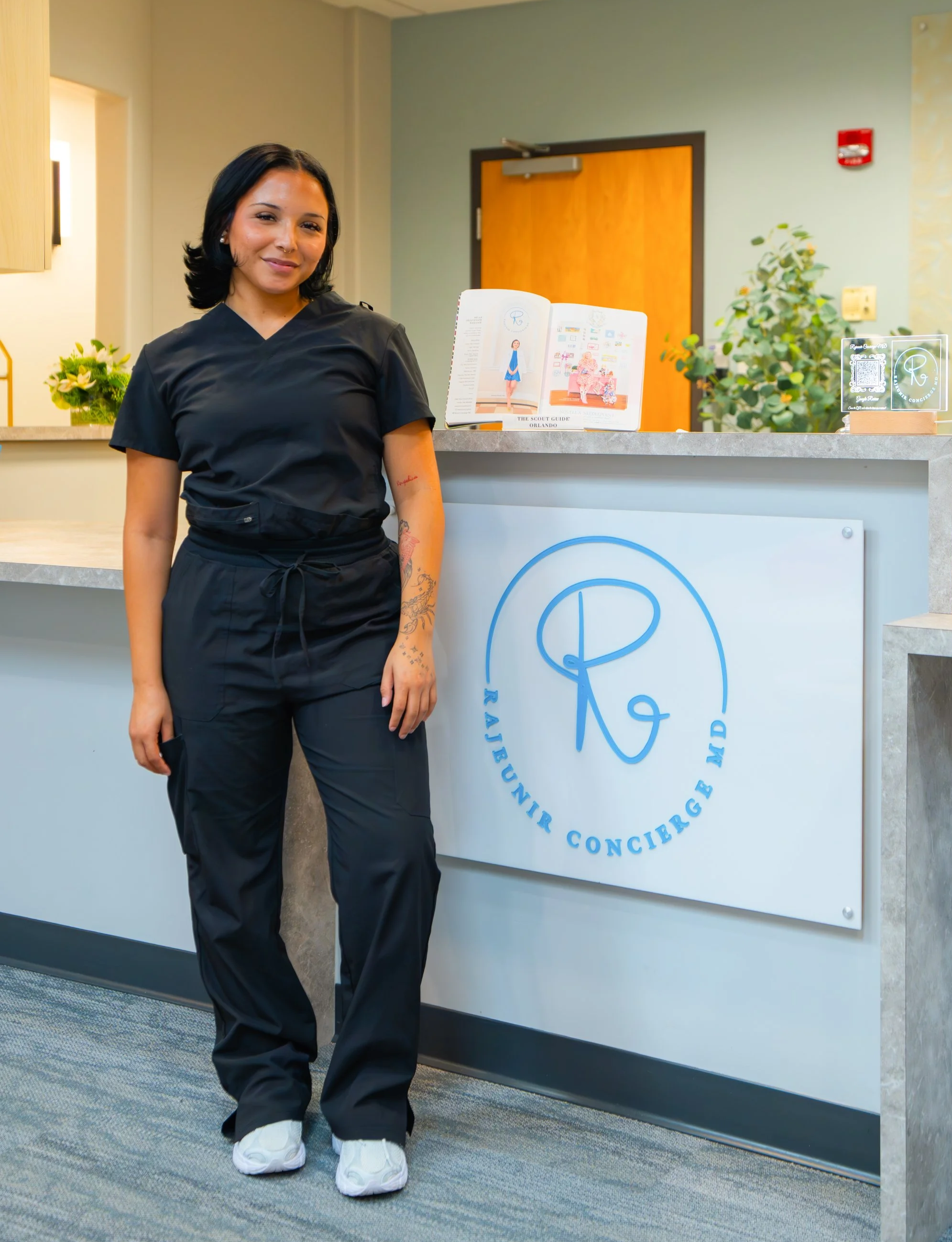 A woman with long brown hair and makeup, wearing a black T-shirt and black gloves, crossing her arms in front of a plain white wall with some green leaves in the foreground.