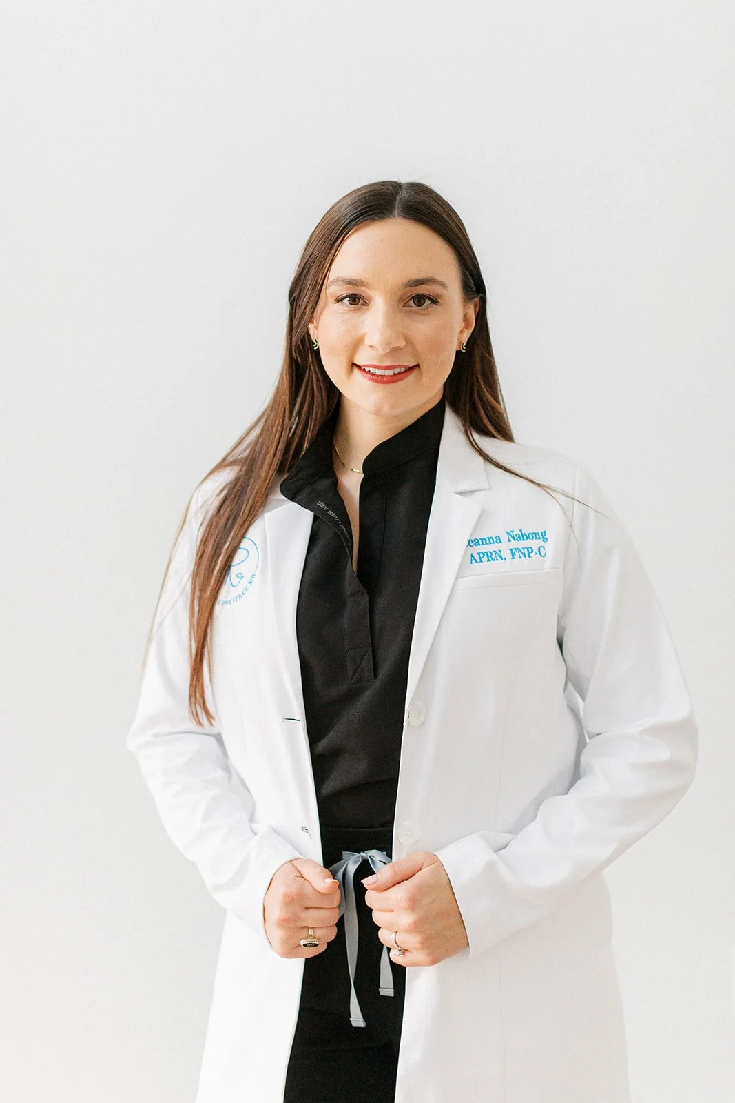 A woman in a white medical coat smiling, with dark hair and a black shirt underneath, standing against a plain white background.