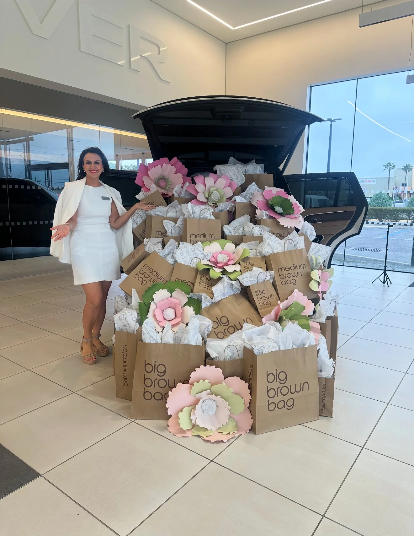 A woman in a white dress standing next to a large car trunk filled with shopping bags and paper flowers, inside a modern showroom with large windows.
