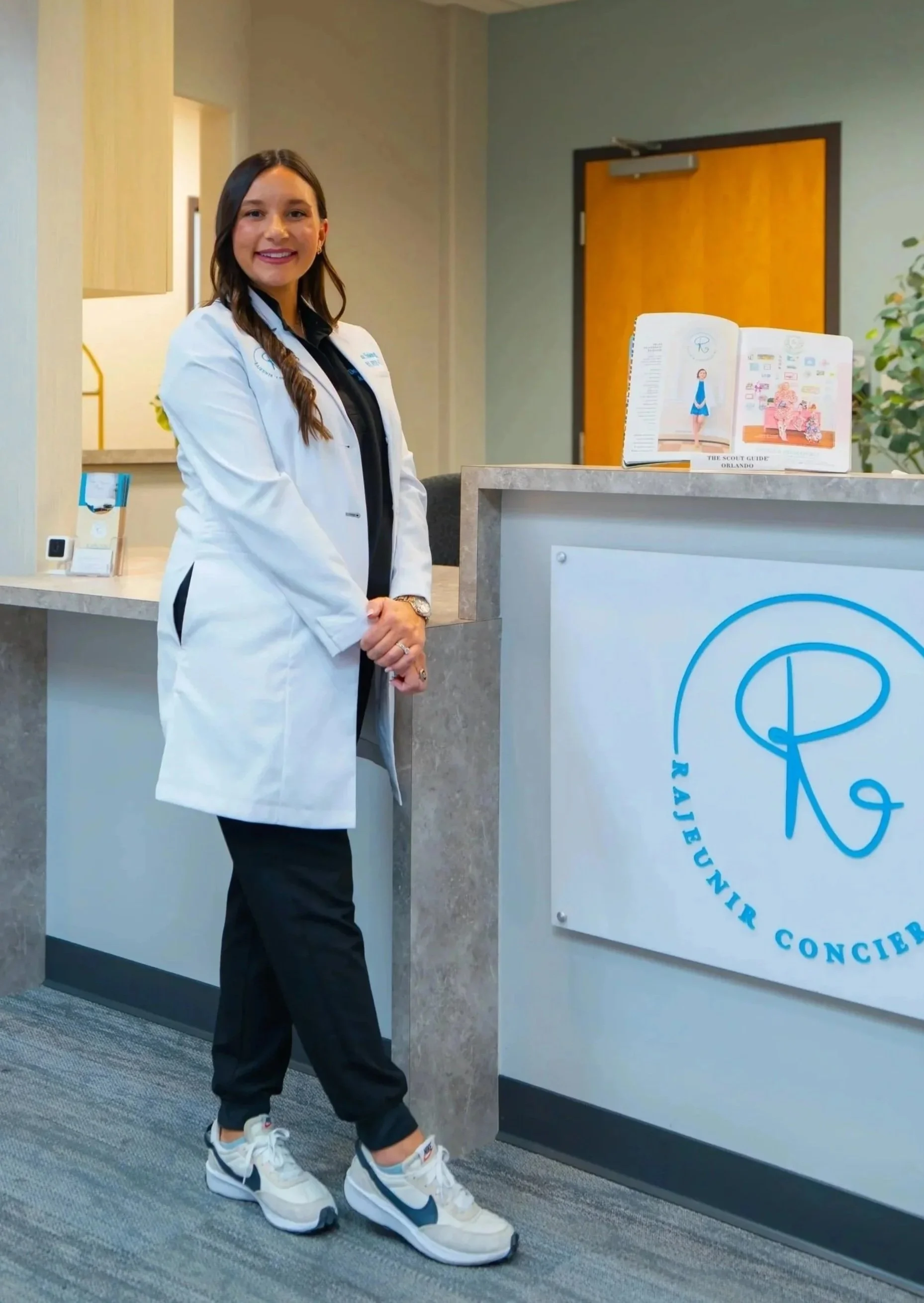 A woman in a white medical coat smiling, with dark hair and a black shirt underneath, standing against a plain white background.