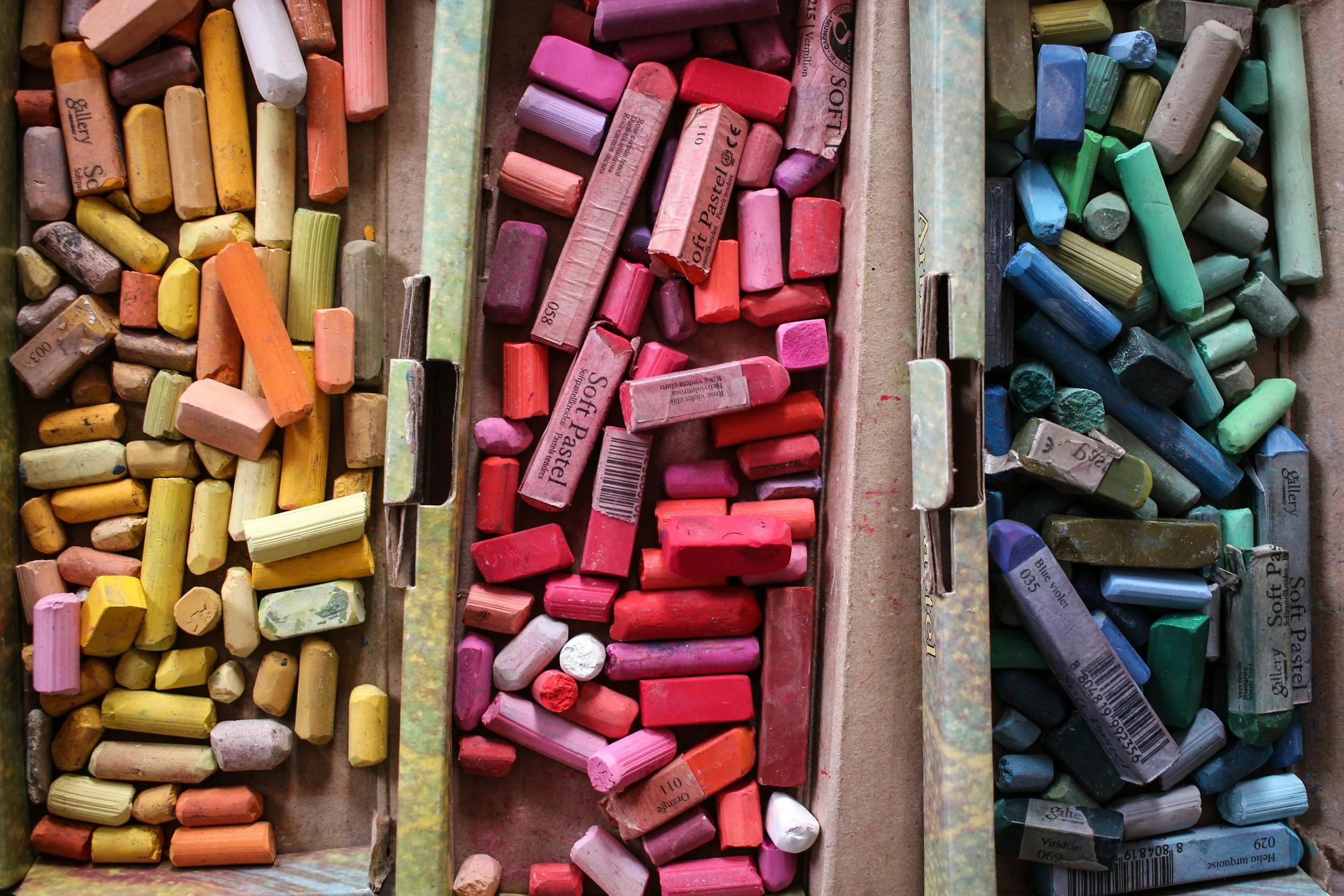 A close up shot of brightly coloured oil pastels, in three colour themed sections. The first is yellow and orange, then there is a pink and red section, and finally a blue and green section.