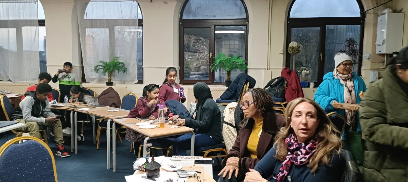 People of a variety of ages are sat around tables in a community space. They are making art together