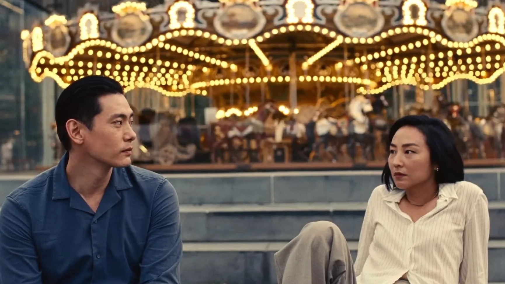 The characters of the film Past Lives look at each other while sitting in front of a merry-go-round