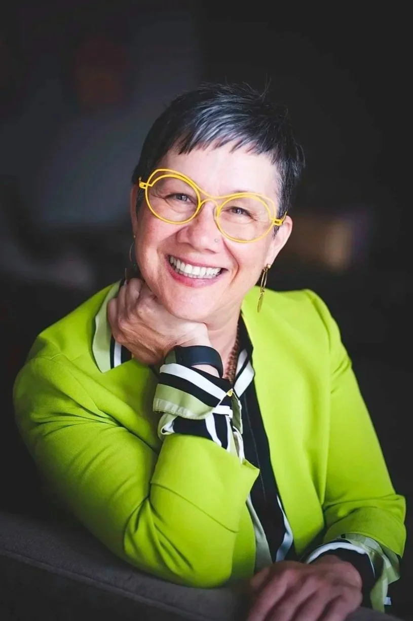 A woman with short dark hair and green eyes wearing yellow glasses, a nose piercing, and a lime green jacket over a dark top, smiling.