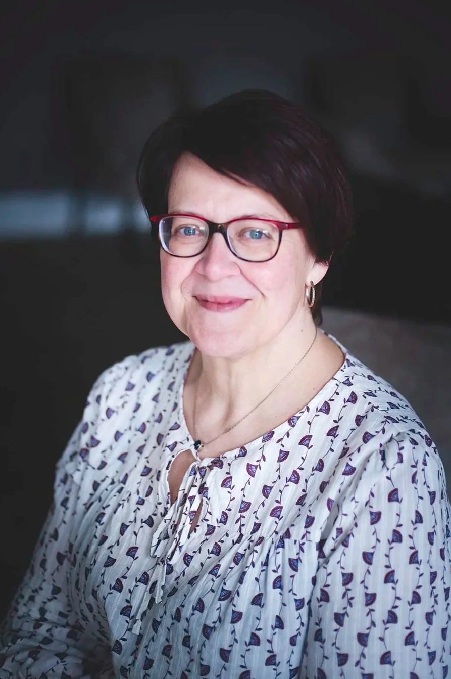 A smiling woman with short dark hair and red glasses standing outdoors with greenery in the background.