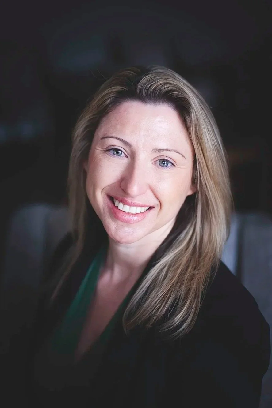 A woman with shoulder-length blonde hair smiling outdoors with a background of green foliage.