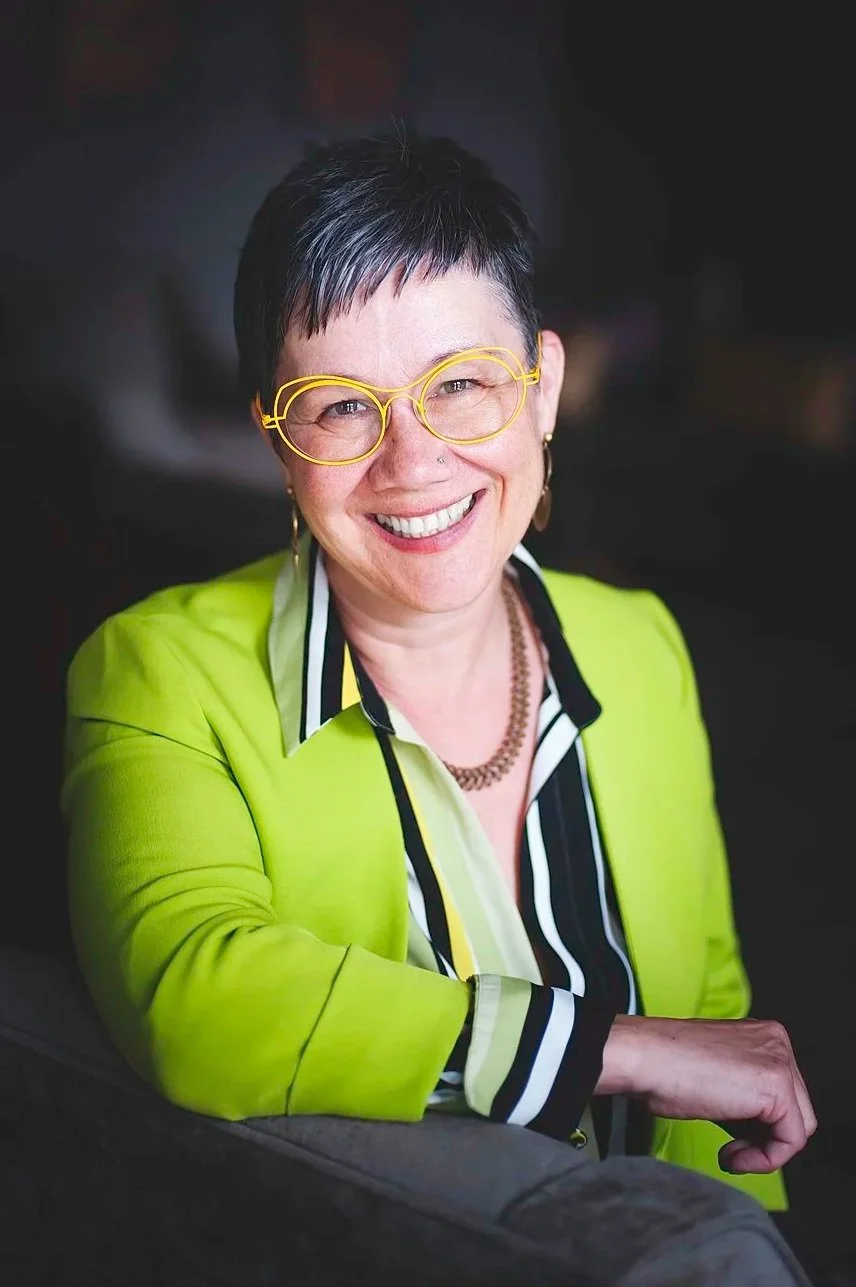 A woman with short dark hair and green eyes wearing yellow glasses, a nose piercing, and a lime green jacket over a dark top, smiling.