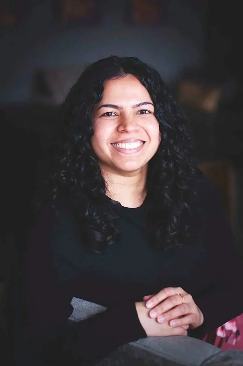 A woman with dark wavy hair smiling, wearing a brown blazer over a black top, sitting near a window with curtains.