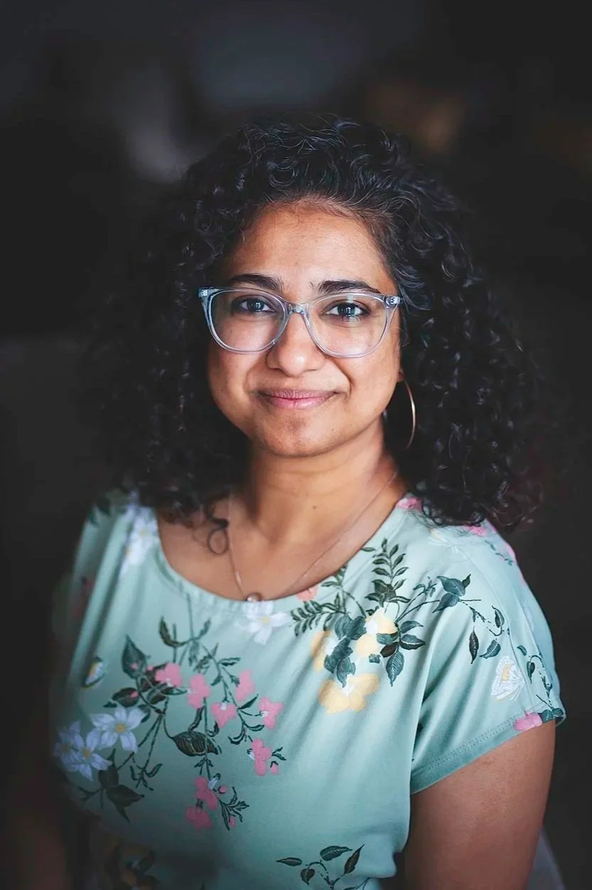 A woman with curly hair and a warm smile, wearing a red top with white dots and a black cardigan, indoors near a window with natural light.