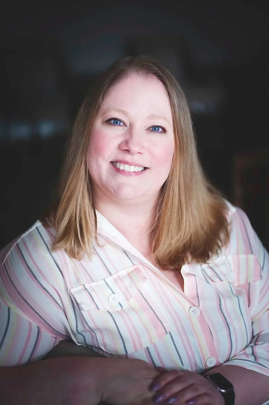 A young woman with wavy brown hair and blue eyes smiling, wearing a floral top, a delicate necklace, and natural makeup, standing against a wooden background.