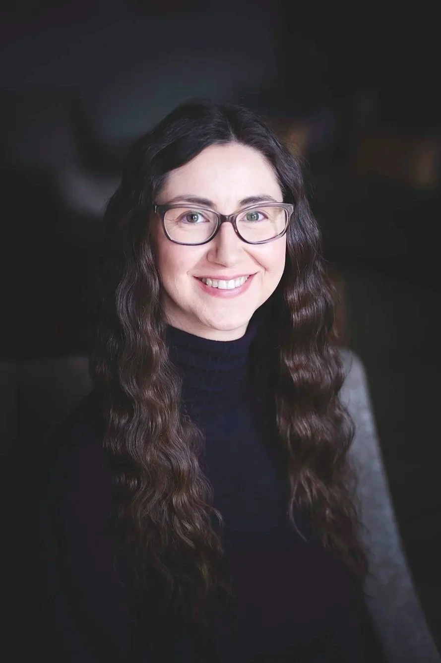 A woman with long brown hair, glasses, and wearing a navy blue top, smiling at the camera in a well-lit room.