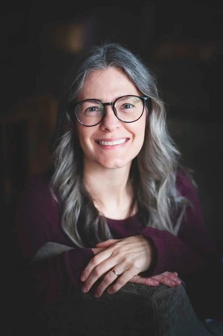 A smiling woman with shoulder-length, grayish hair, wearing glasses and a maroon top, in a cozy home interior.