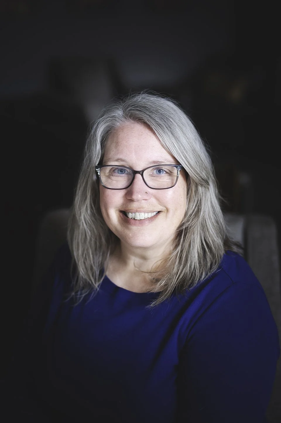 A woman with long gray hair and glasses smiling outdoors with blurred green trees and grass in the background.