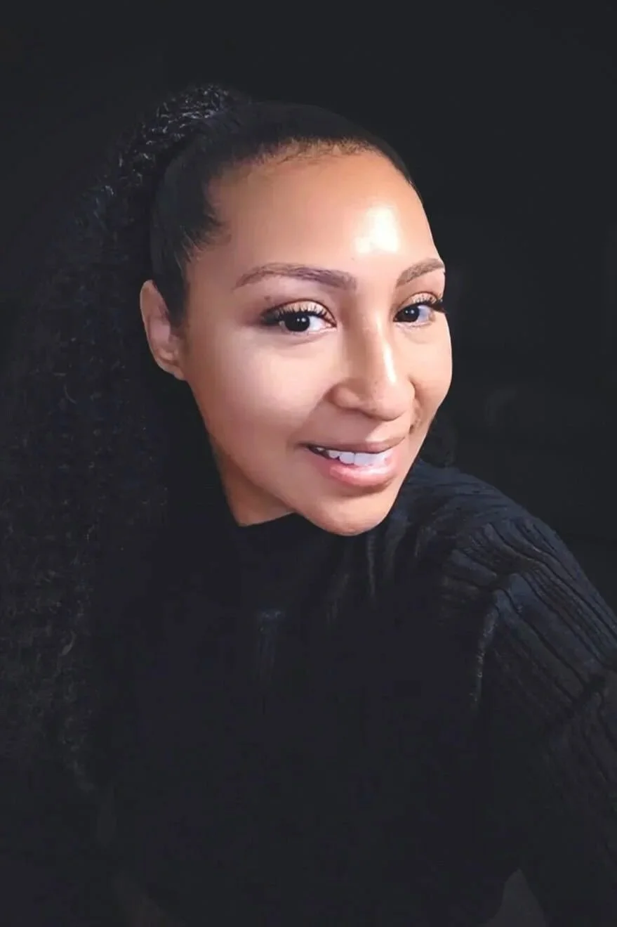 A smiling woman with curly hair styled in a high ponytail, wearing a black top, sitting in front of a gray background.