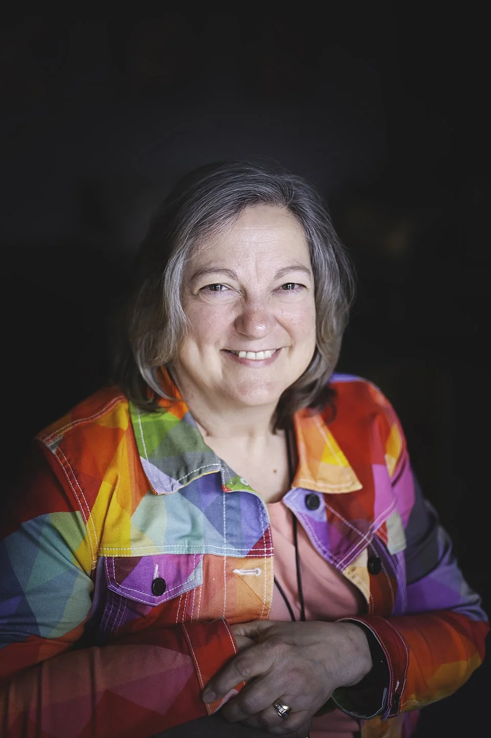 A smiling woman with shoulder-length brown hair, wearing a green jacket and a patterned scarf, posed against a dark background.
