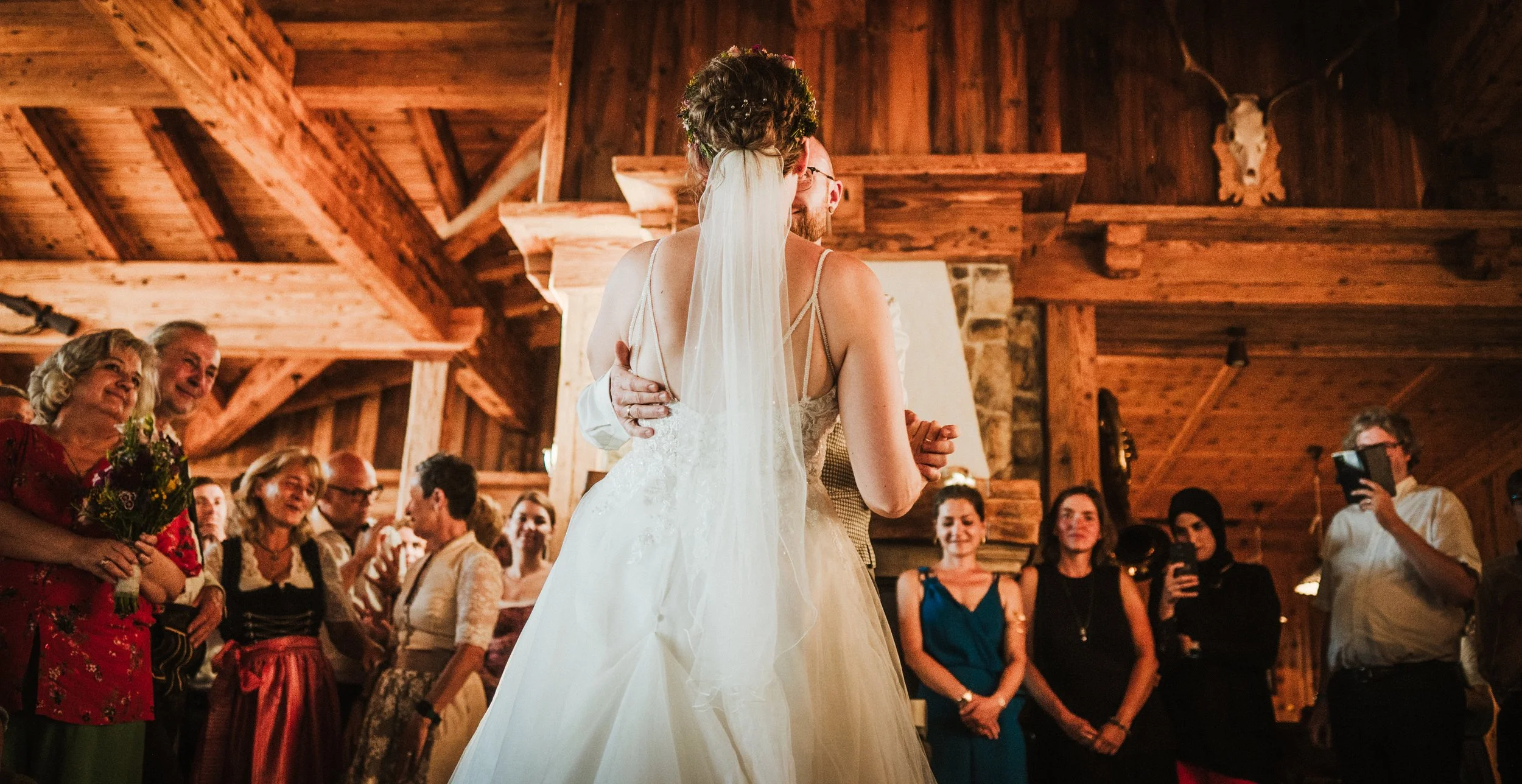 Bride and groom dancing at wedding reception with guests watching in rustic wooden venue.