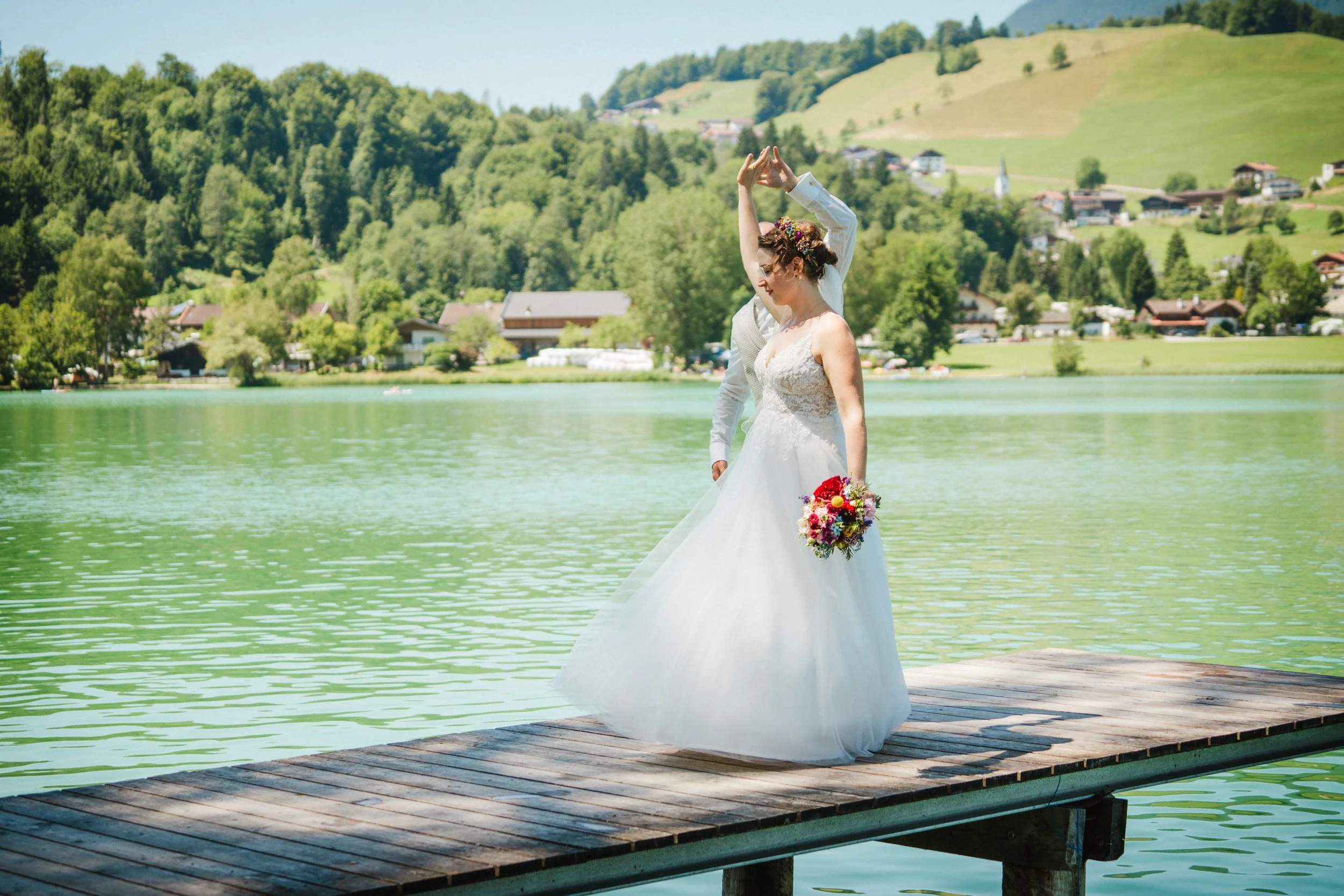 Bride and groom dancing on a dock by a lake, surrounded by wooded hills.
