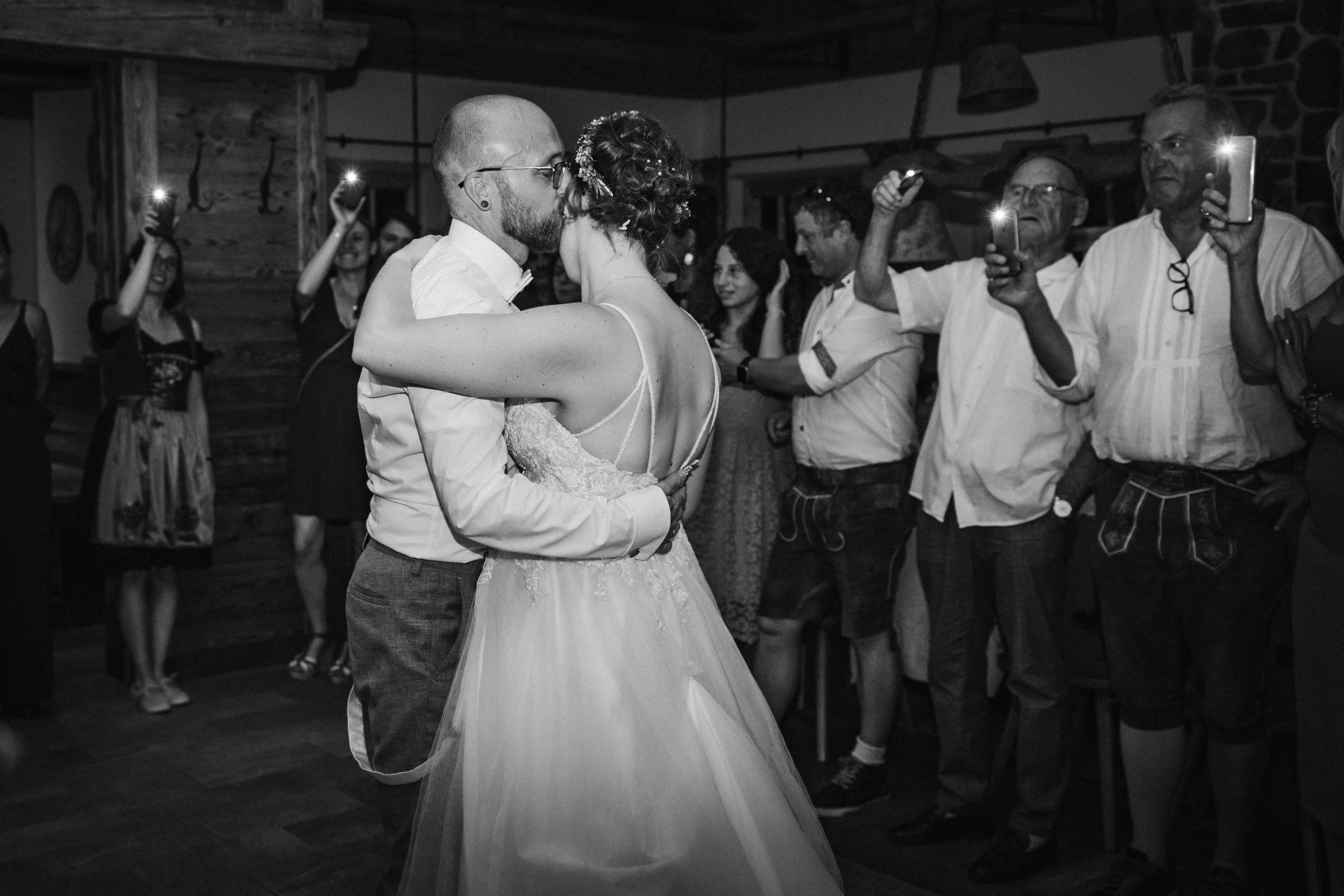 Bride and groom kiss at wedding with guests taking photos, black and white.