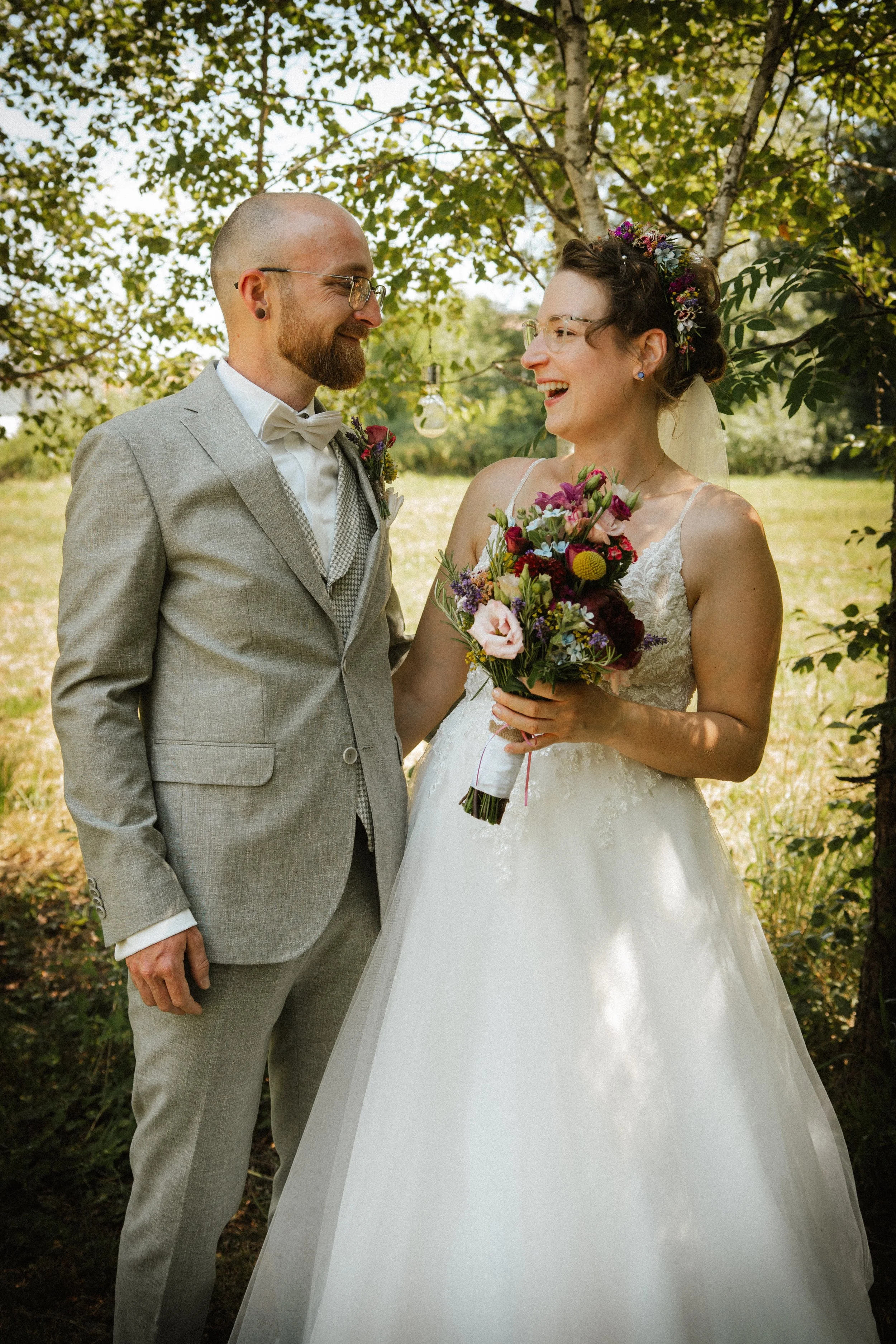 Bride and groom smiling, outdoors, bride holding bouquet, groom in grey suit.