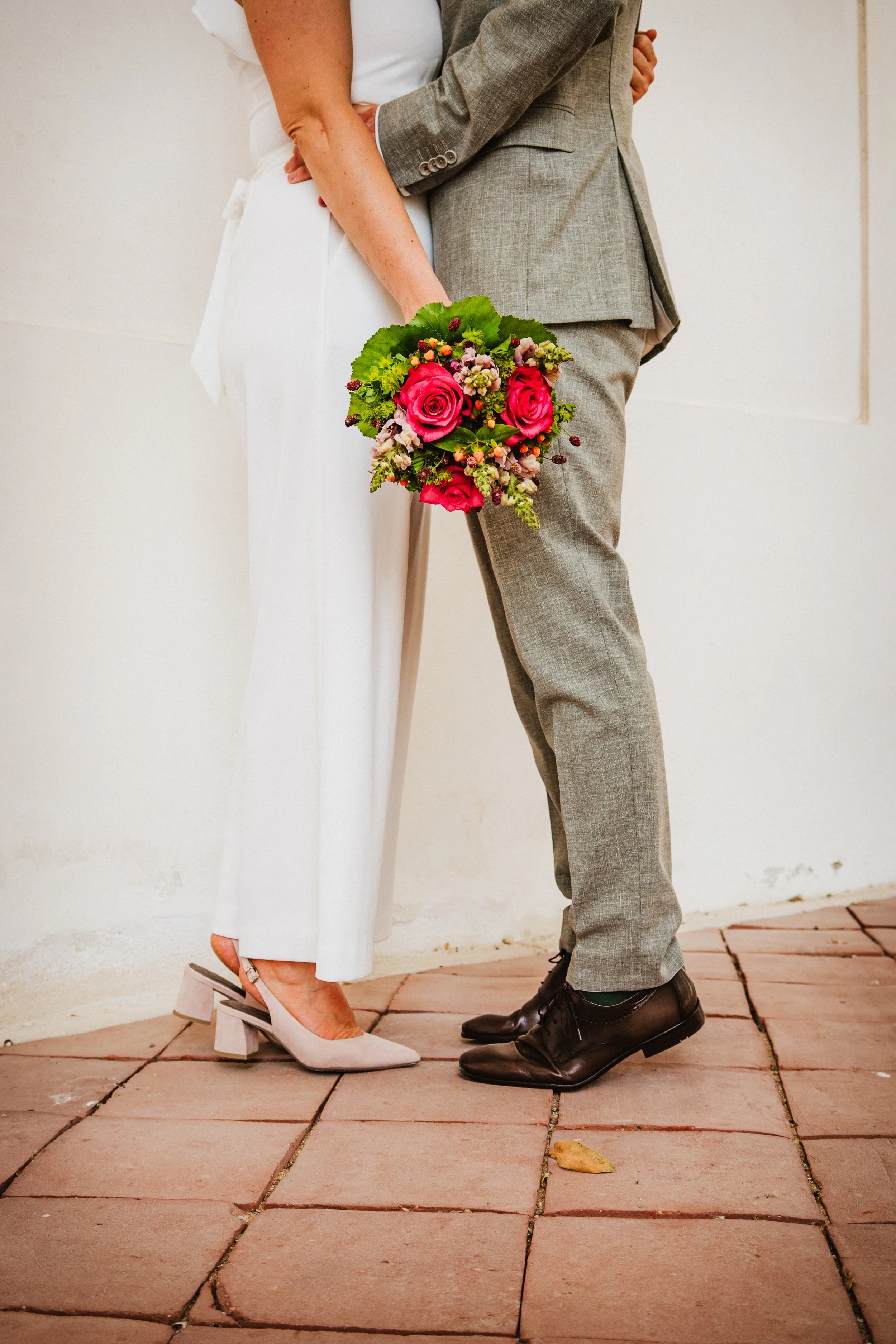 Close-up of a couple in wedding attire, woman holding a bouquet of pink roses, standing on a tiled surface.