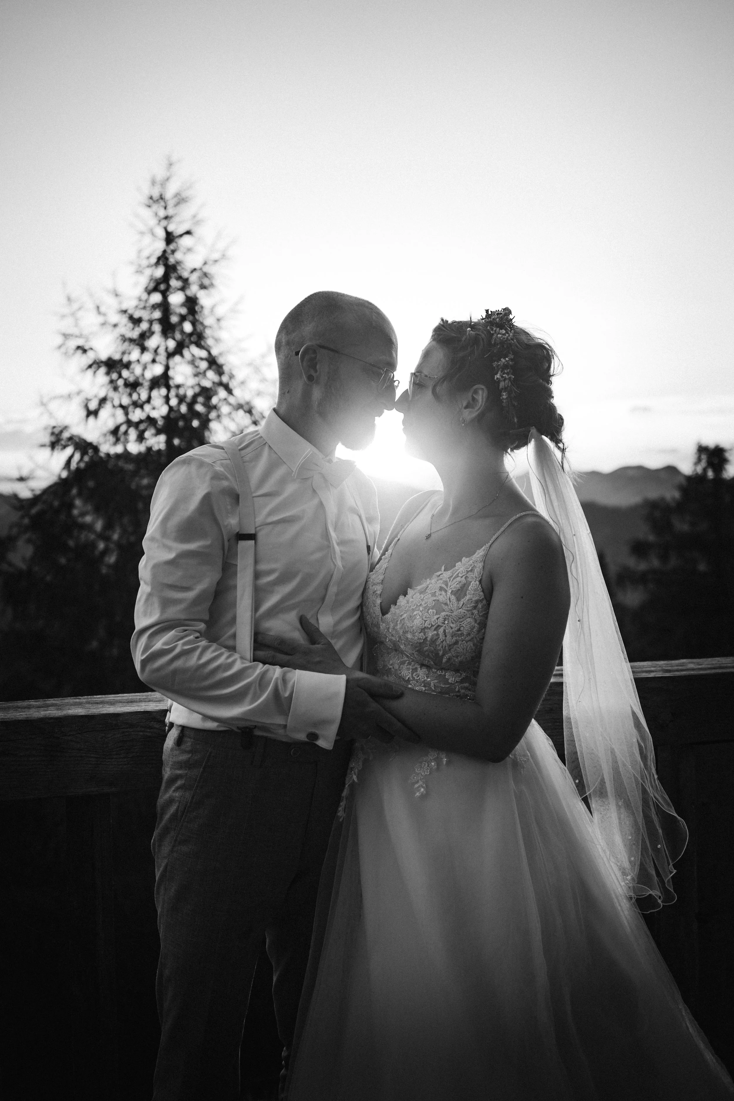 Silhouette of a couple in wedding attire embracing, with a scenic background of trees and sunset.