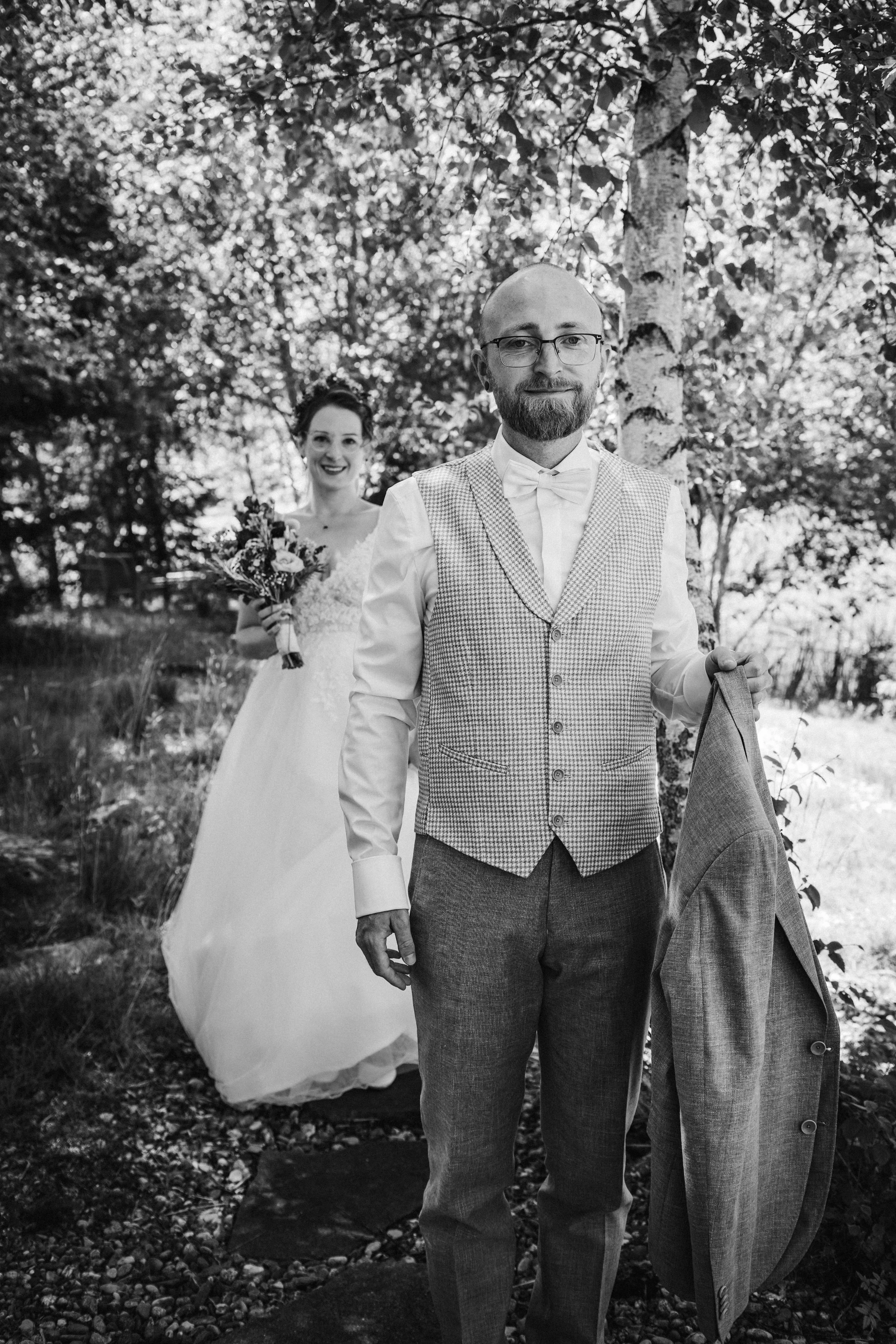 Black and white photo of a bride and groom in a garden, groom holding jacket and bride holding flowers, both smiling.