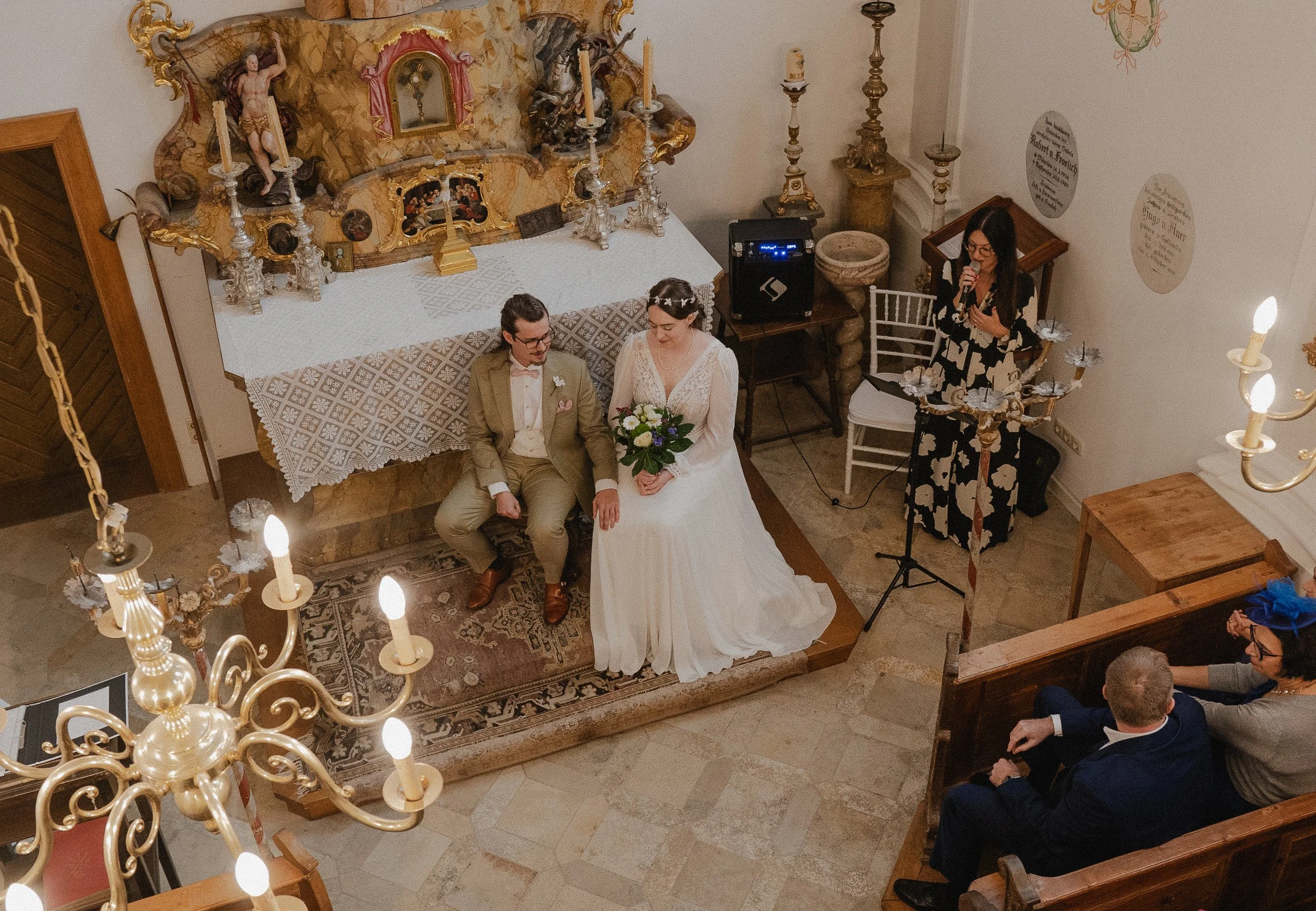 Wedding ceremony in a church with a bride and groom seated near an altar, surrounded by ornate decorations and a chandelier above; a woman singing or speaking nearby.