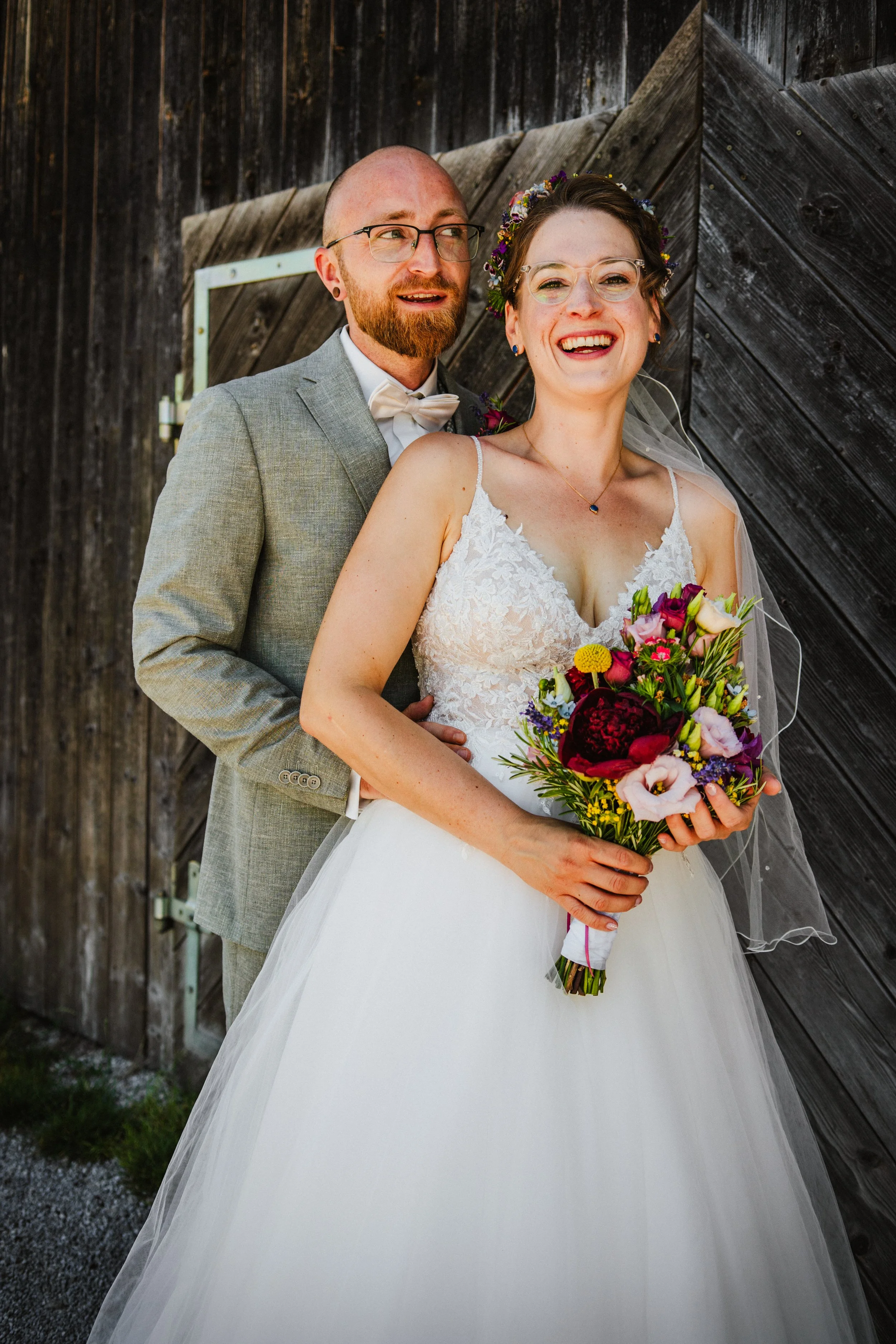 Bride and groom pose happily at wedding with bouquet, rustic background.