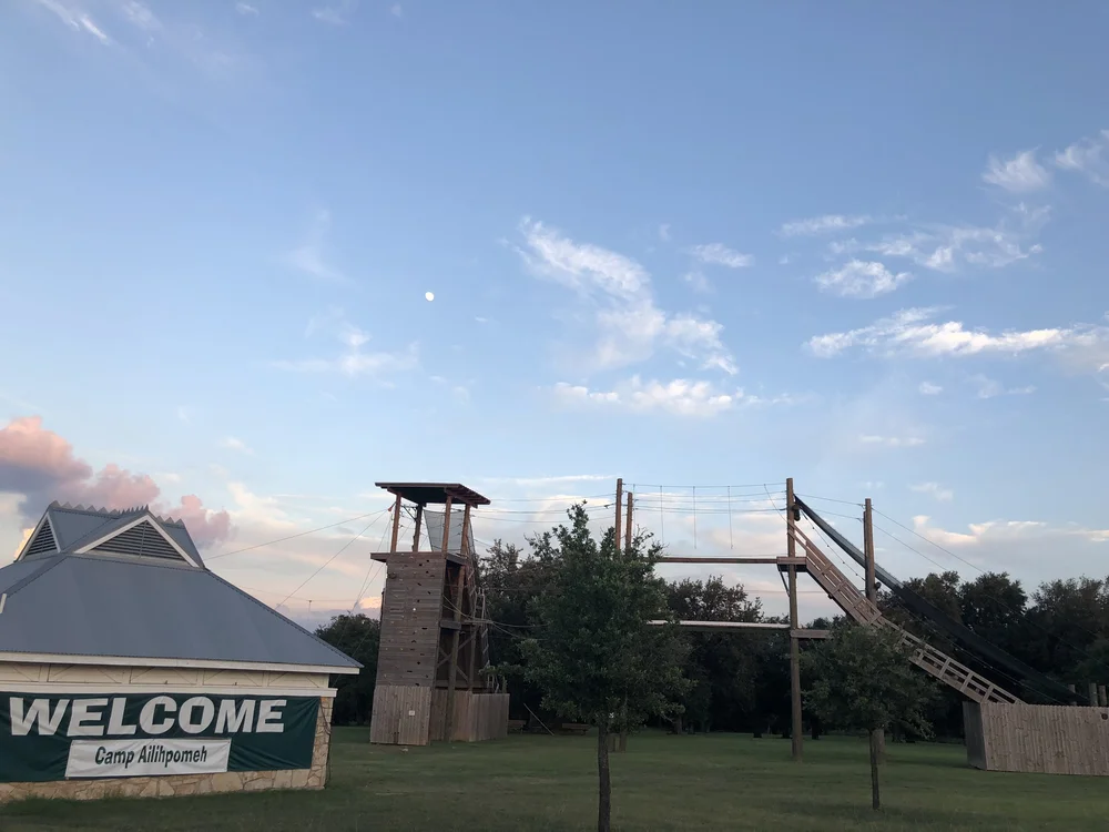 A sign welcoming campers to Camp Ailihpomeh, with the ropes course behind it