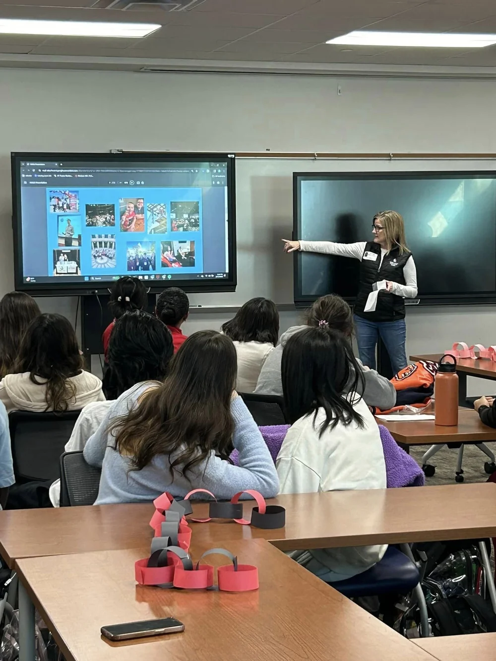 Students seated facing a Lone Star staff member who is presenting at a classroom screen, with a red and gray paper chain lying on the table in the foreground