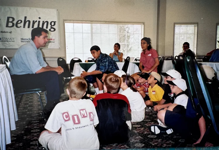 Children seated on the floor at a chapter education session, a sponsor banner on the wall behind them