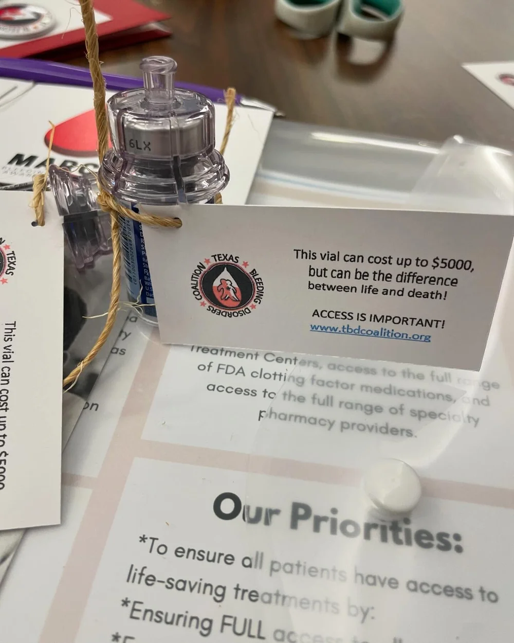 A clotting factor vial with a tag reading: this vial can cost up to $5,000, but can be the difference between life and death. Access is important. It rests on the Coalition's priorities handout.