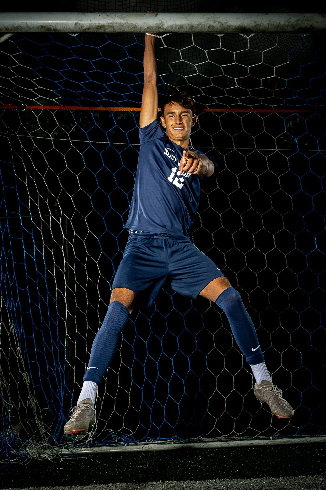 A young male soccer player in a navy blue uniform hanging from a goalpost inside a soccer net, pointing and smiling at the camera.