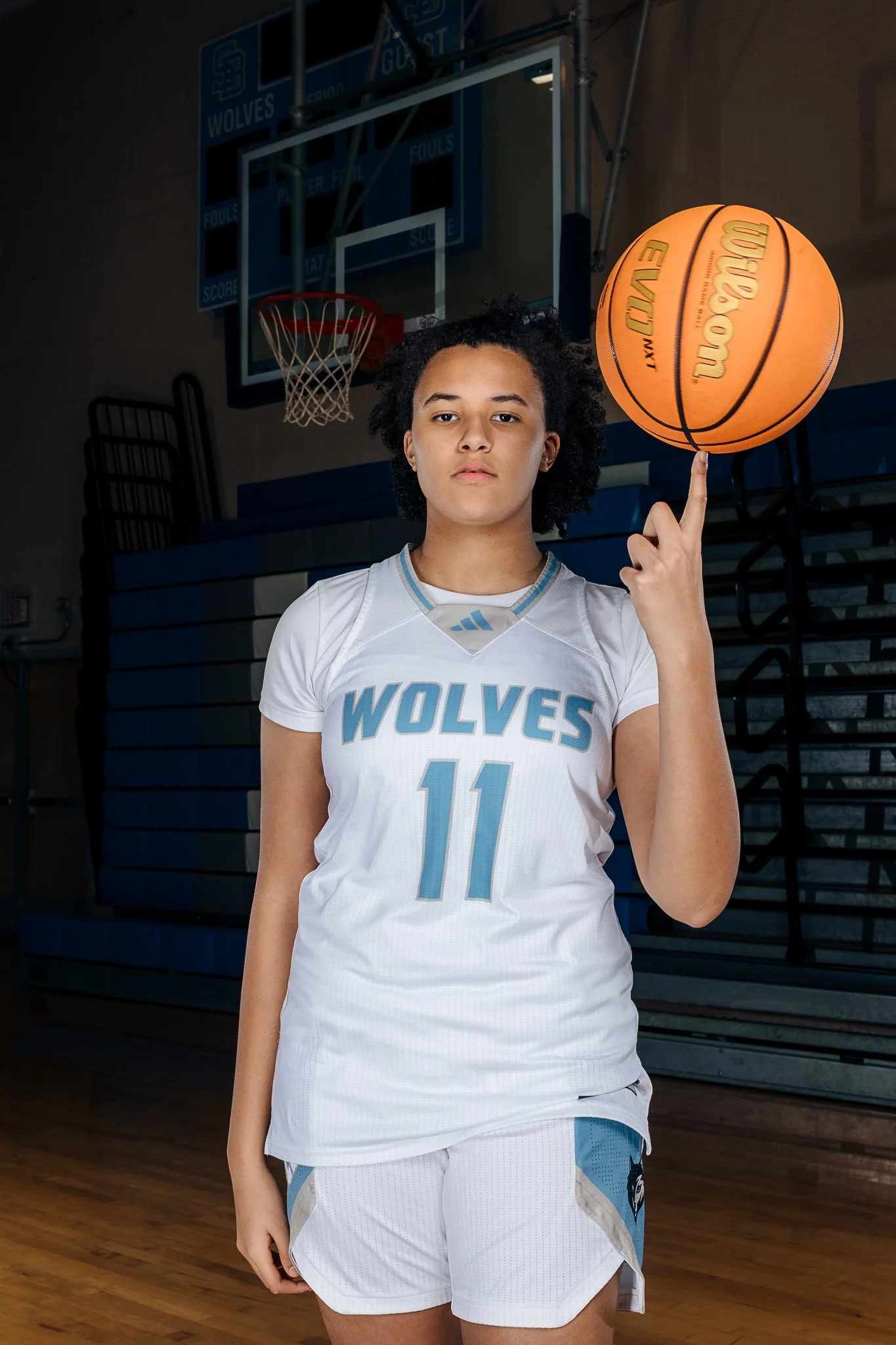 A young female basketball player in a white uniform with blue letters and the number 11, spinning a basketball on her finger in a gymnasium.