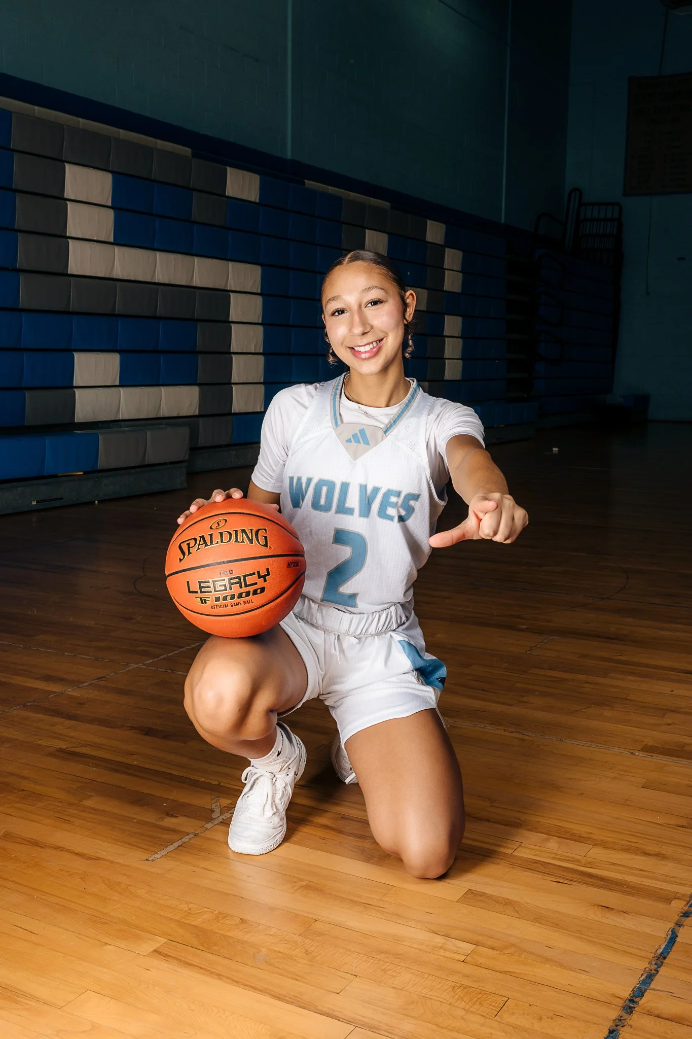 A young female basketball player in a white and blue Wolves uniform kneeling on a basketball court, holding a basketball and pointing towards the camera, smiling.