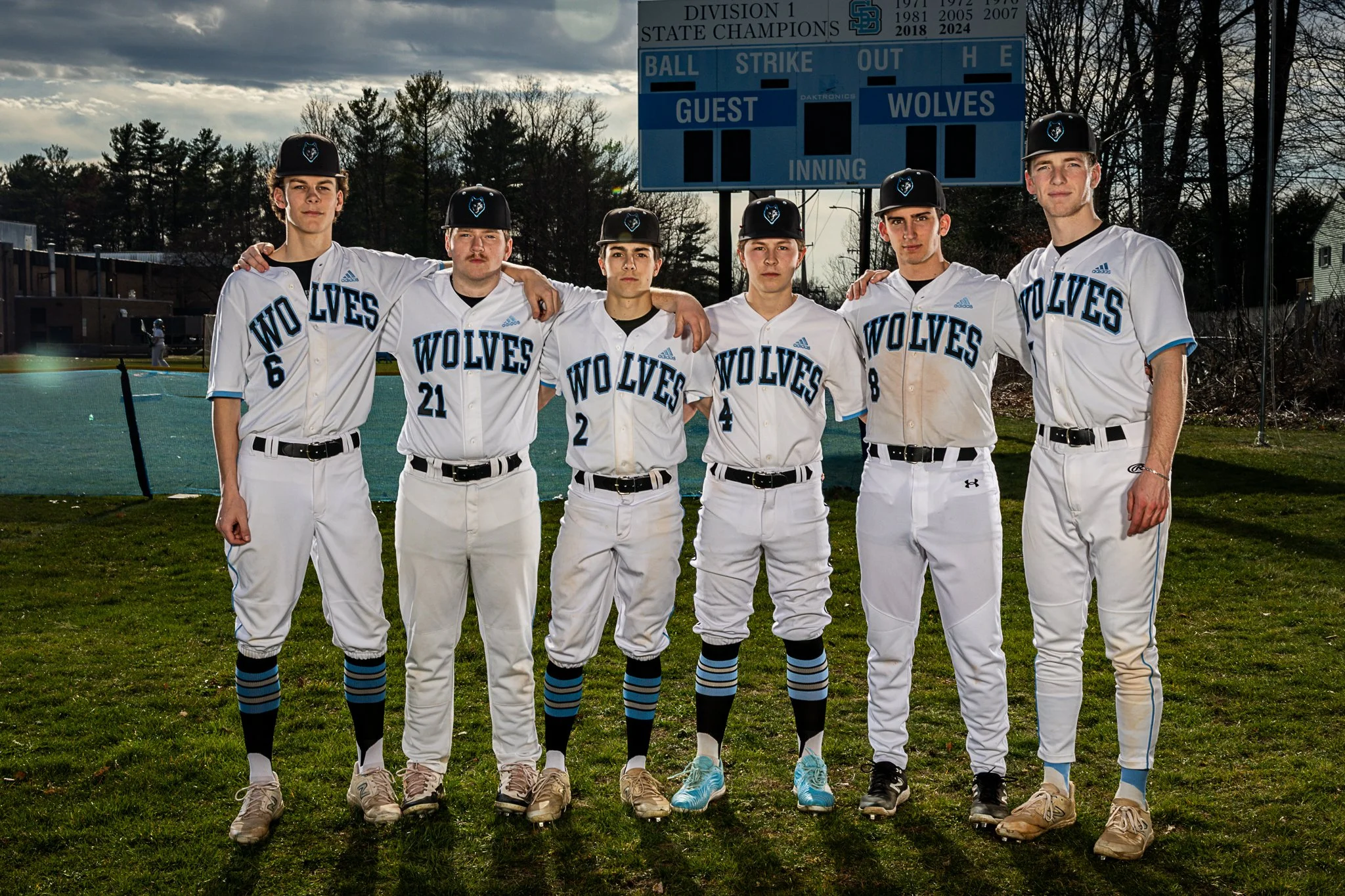 Six baseball players in white uniforms with blue and black accents standing on a field with their arms around each other's shoulders. They are wearing black hats with a logo and are standing in front of a scoreboard, with trees and a cloudy sky in th