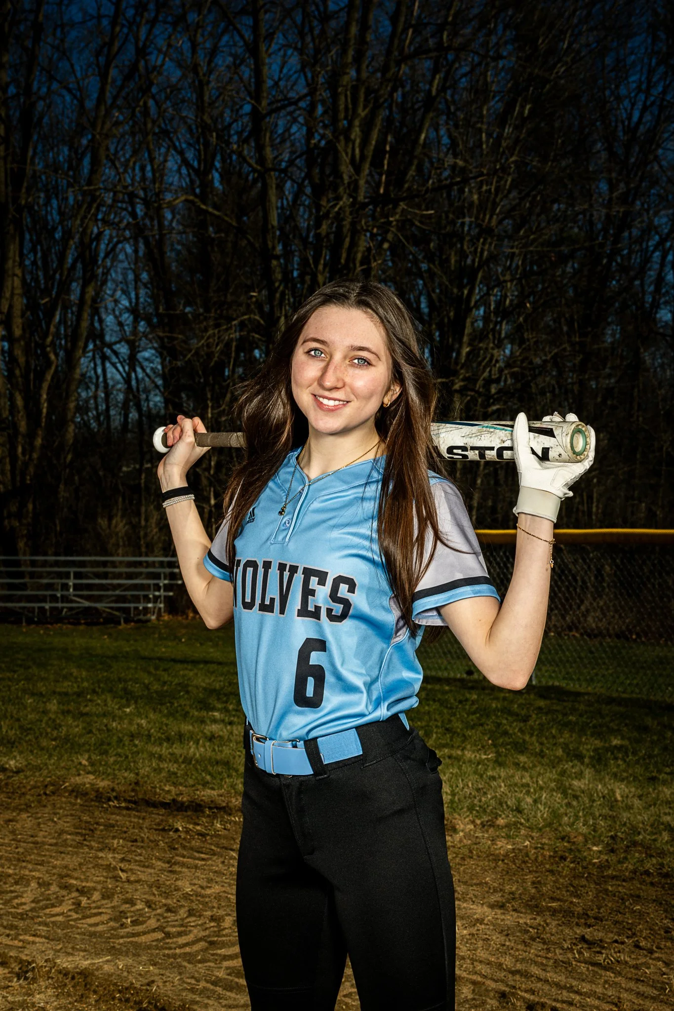 A young woman in a light blue softball uniform with 'WOLVES' and the number 6 on the front, holding a baseball bat over her shoulders, standing on a baseball field at dusk with leafless trees in the background.