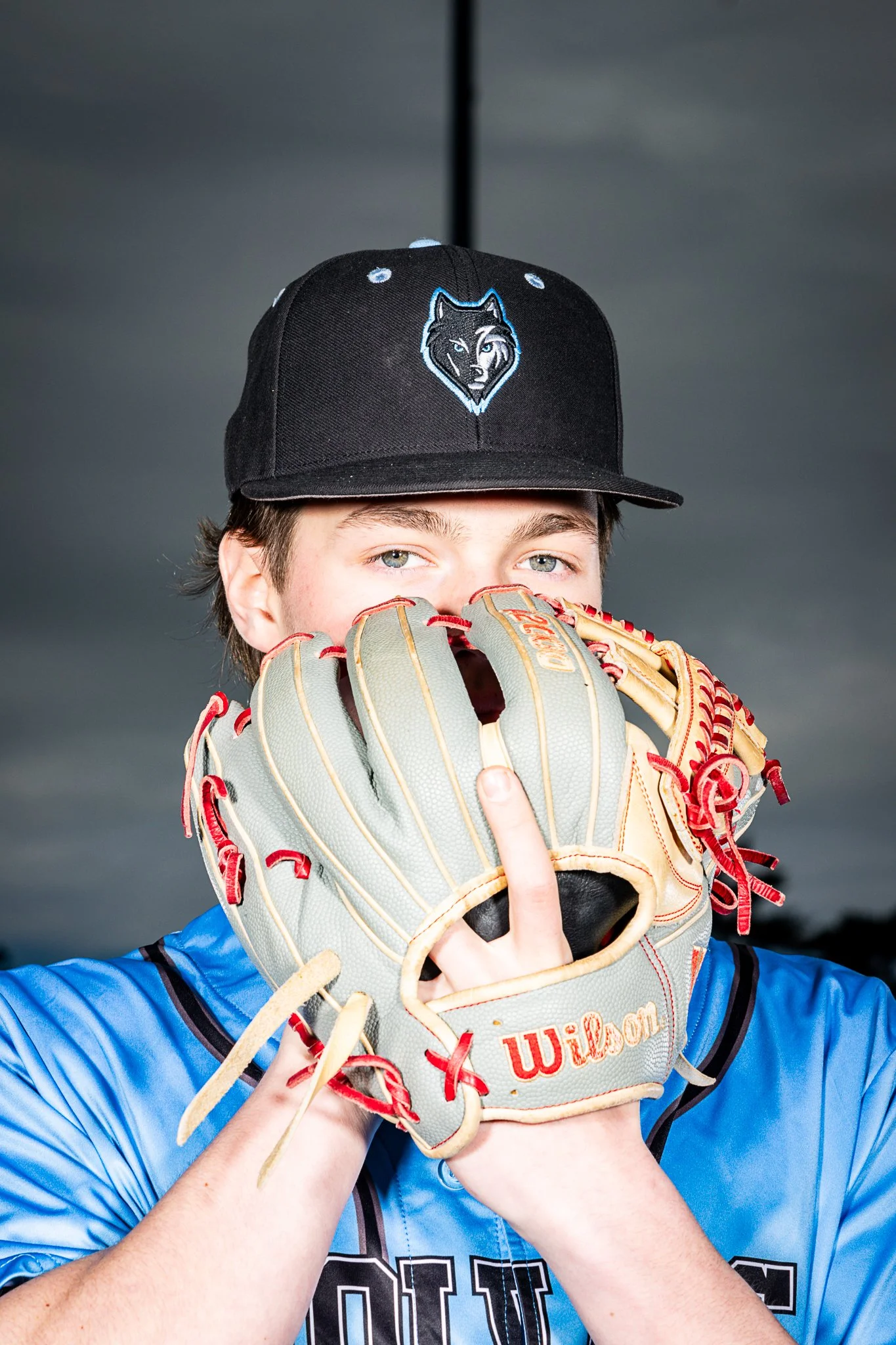 A young baseball player wearing a black cap with a wolf logo, holding a baseball glove up to his face and looking directly at the camera. Dark cloudy sky in the background.