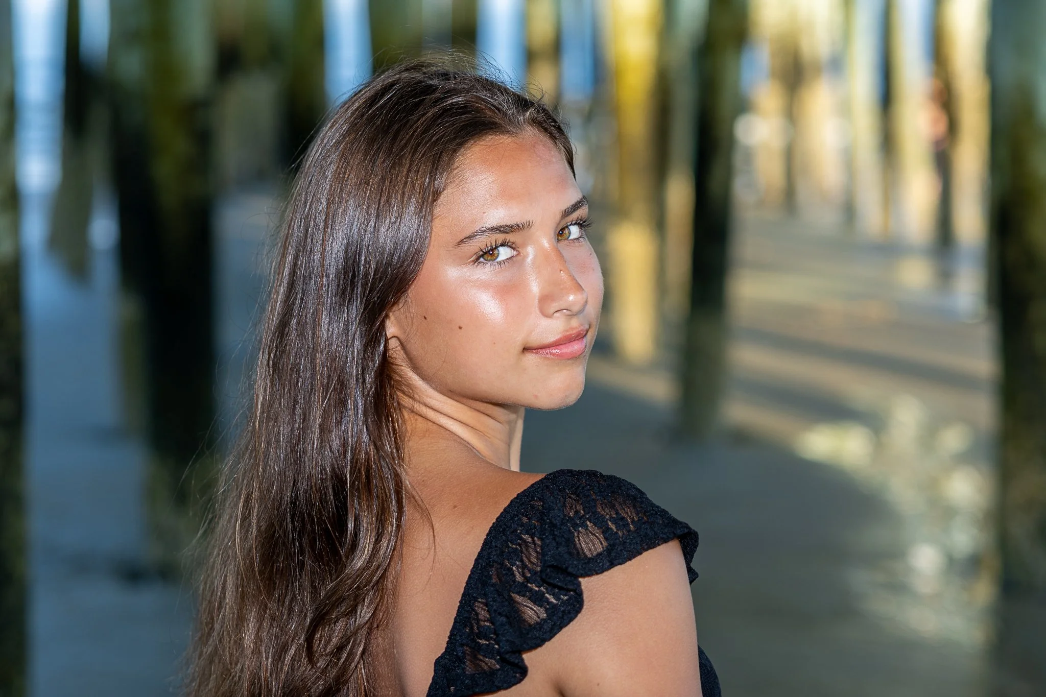 A young woman with long brown hair and a black top outdoors, with trees in the background.