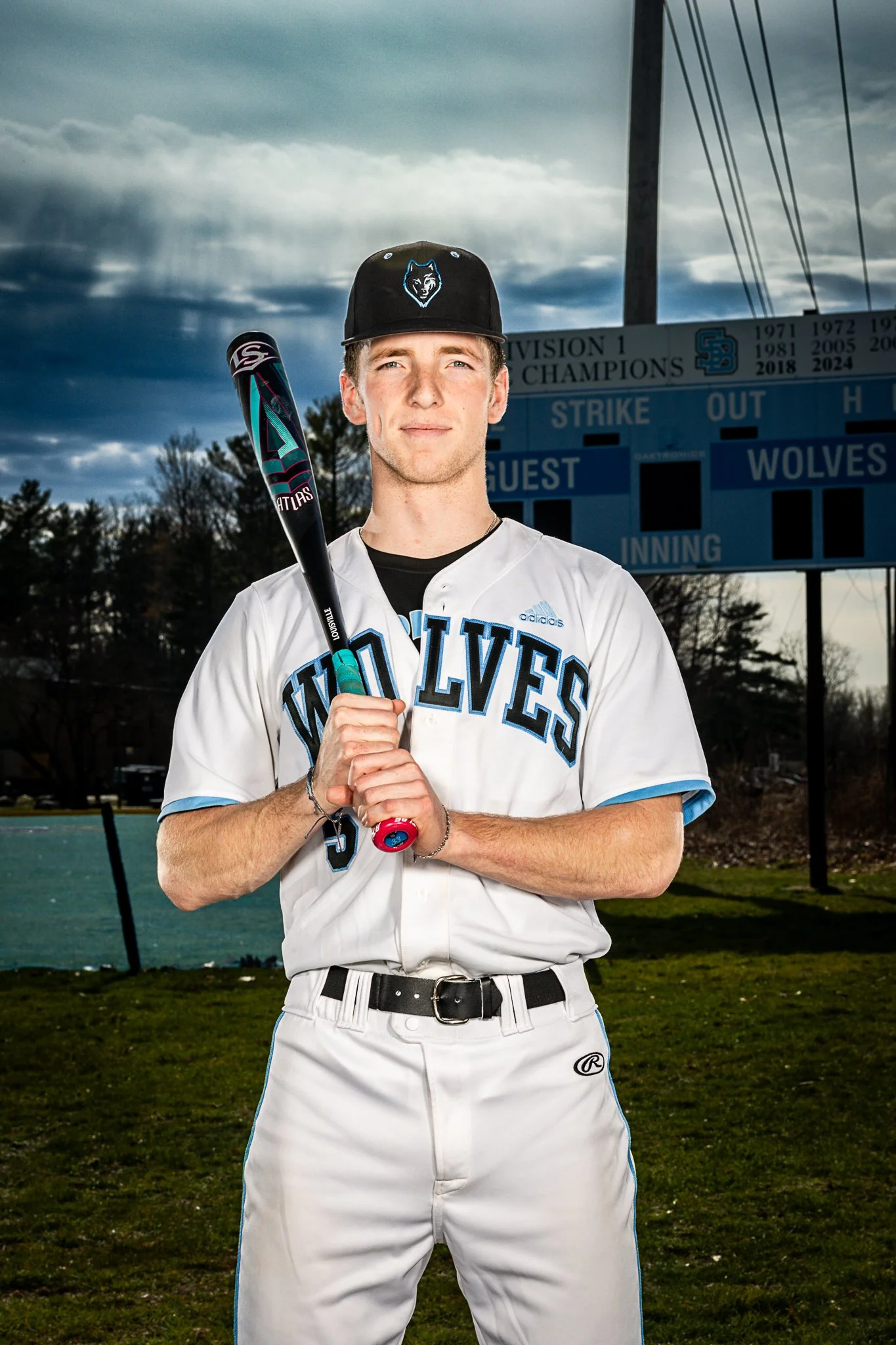 A young man in a white baseball uniform with blue and black accents, holding a bat, standing in front of a baseball scoreboard on a field during cloudy weather.