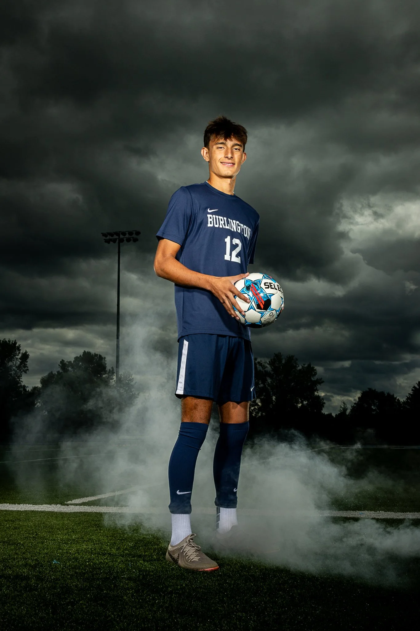 A young male soccer player wearing a blue jersey with the number 12 and 'BURLINGTON' written on it, holding a soccer ball, standing on a field with dark storm clouds overhead and fog around his feet.
